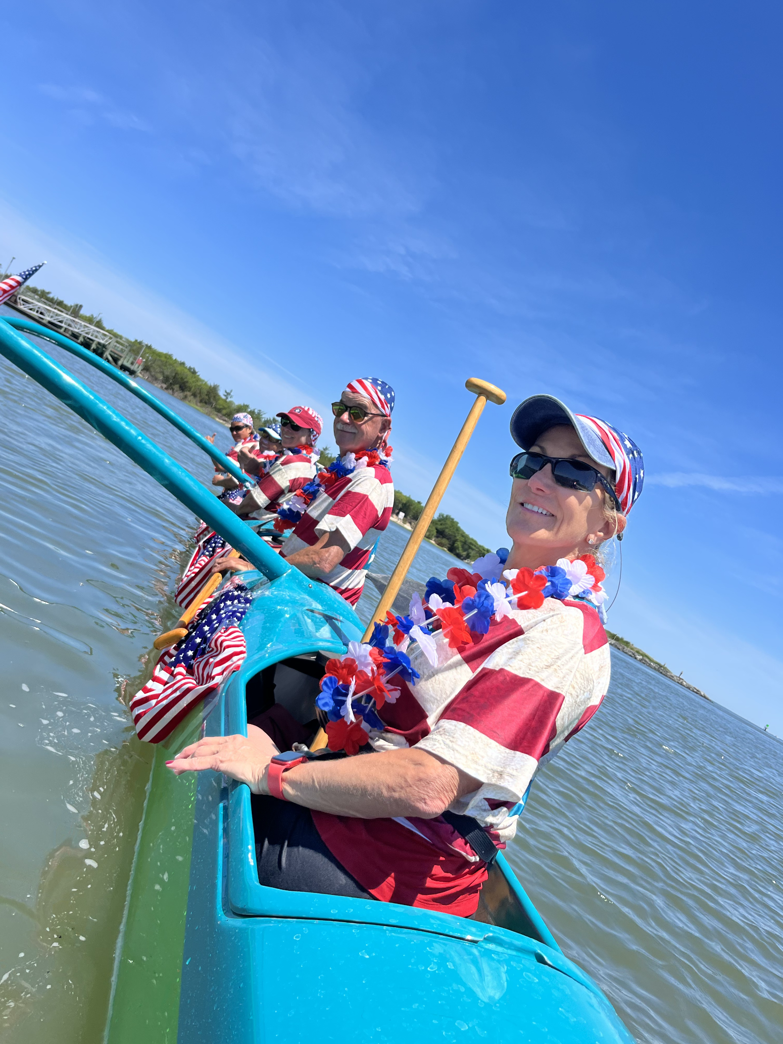 Group of people wearing patriotic American-themed clothing and accessories, paddle boating on a lake on a sunny day.