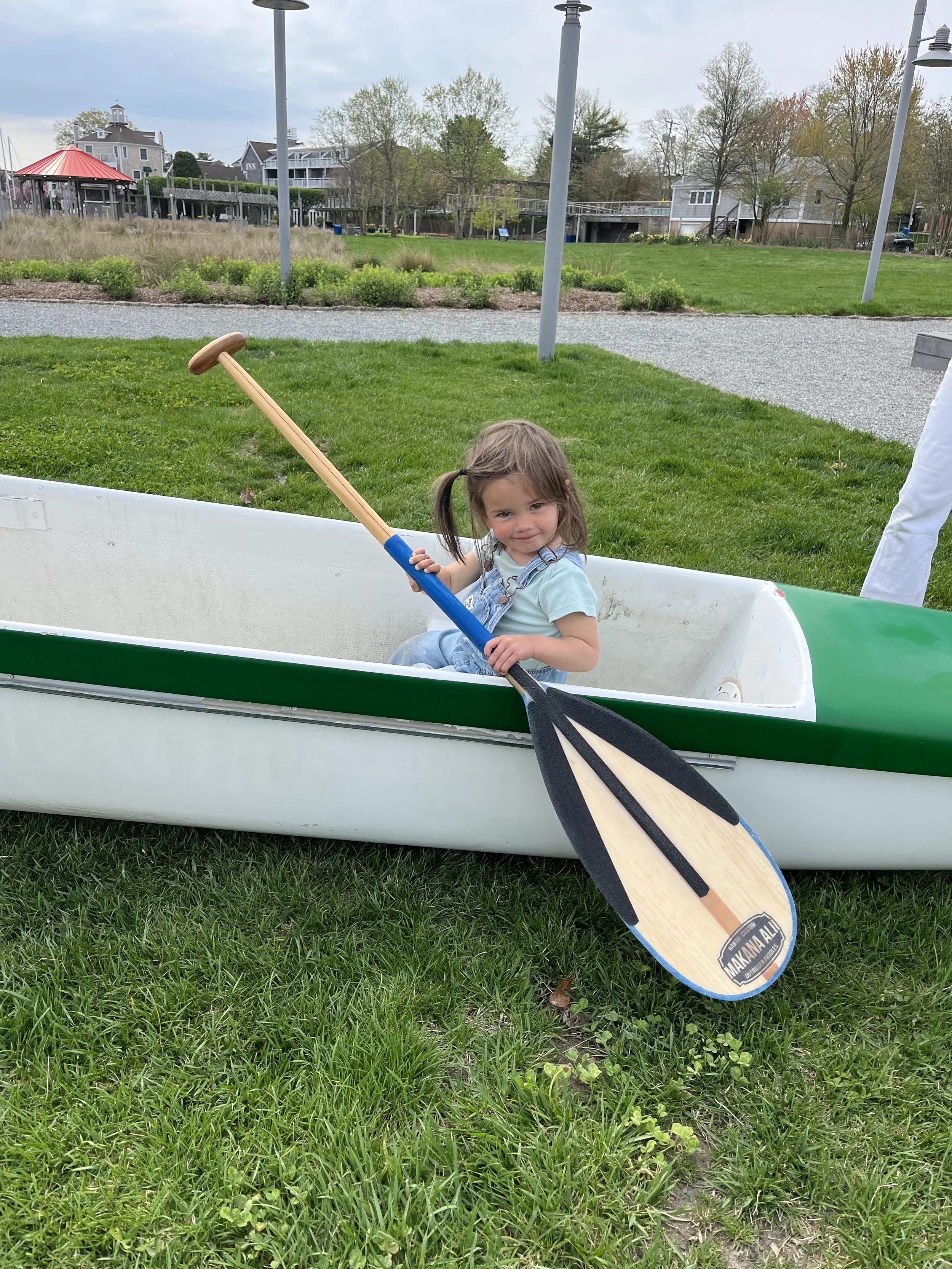 A young girl with brown hair in pigtails sitting in a green and white kayak holding a black and wooden paddle, in a park with grass, trees, and buildings in the background.