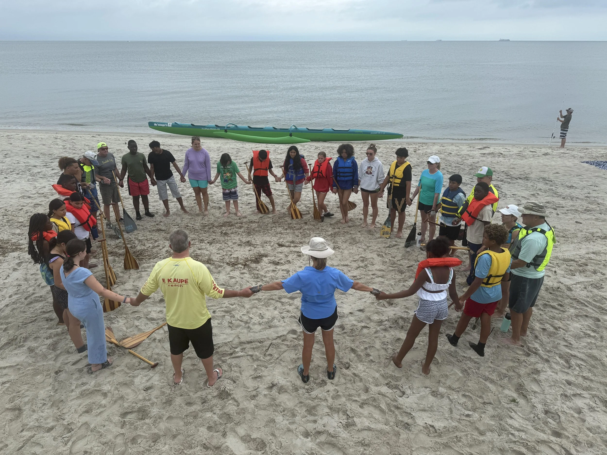 A diverse group of people holding hands in a circle on a sandy beach, with an ocean and ships visible in the background, during daytime. Some participants are wearing life jackets and holding paddles.