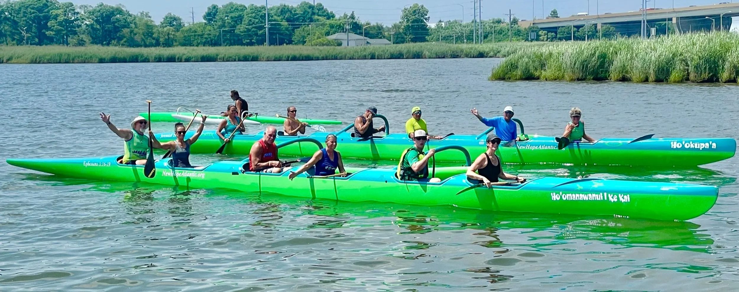 Group of people paddling in two bright green ocean kayaks on a calm body of water during the daytime, with some of the individuals smiling and waving at the camera.