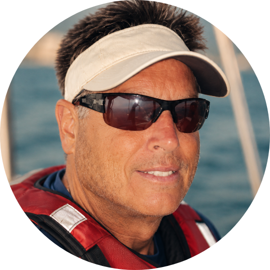 Man wearing sunglasses, a beige visor, and a red life jacket on a boat with water in the background.