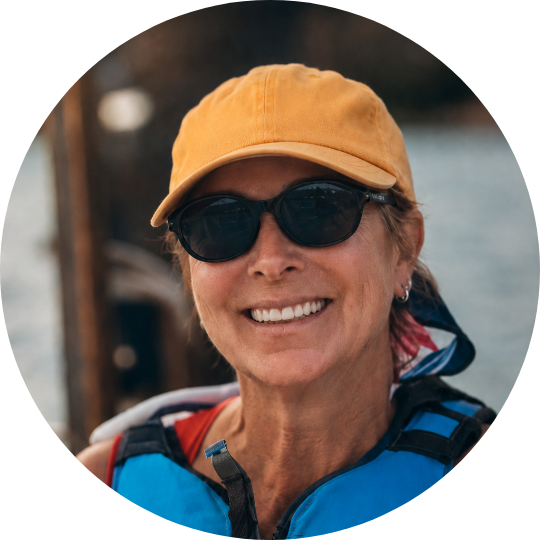 Smiling woman wearing a yellow cap, black sunglasses, and a life jacket outdoors near water.