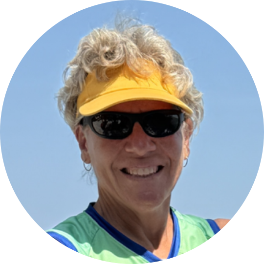 A smiling woman wearing a yellow visor, black sunglasses, earrings, a green athletic top, and a blue sports bra underneath, against a clear blue sky.