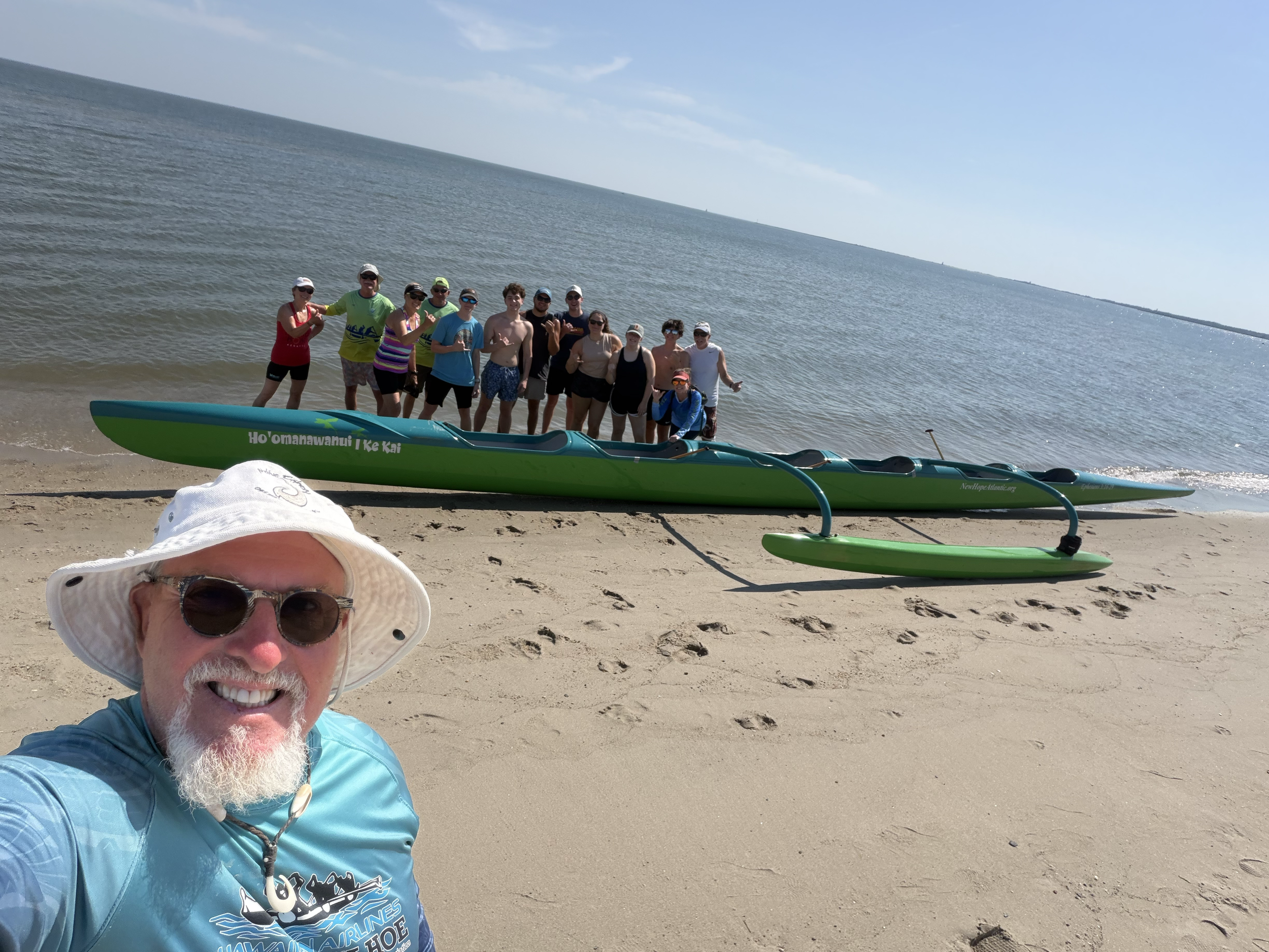 A group of people standing on a beach with a kayak and a paddle in the foreground. The group is smiling and waving, with the ocean and clear sky in the background.
