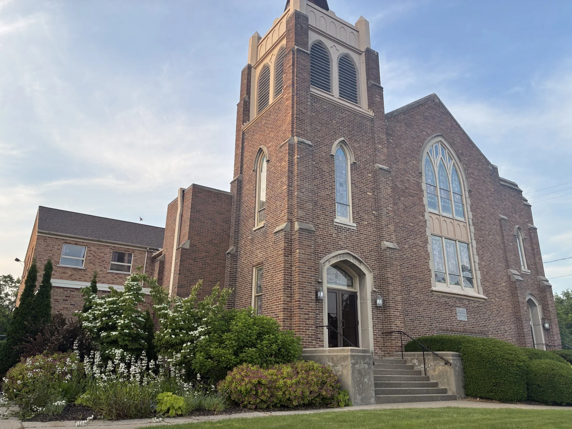 A historic red brick church with bell tower and stained glass windows, surrounded by well-maintained garden and shrubbery, under a clear blue sky.