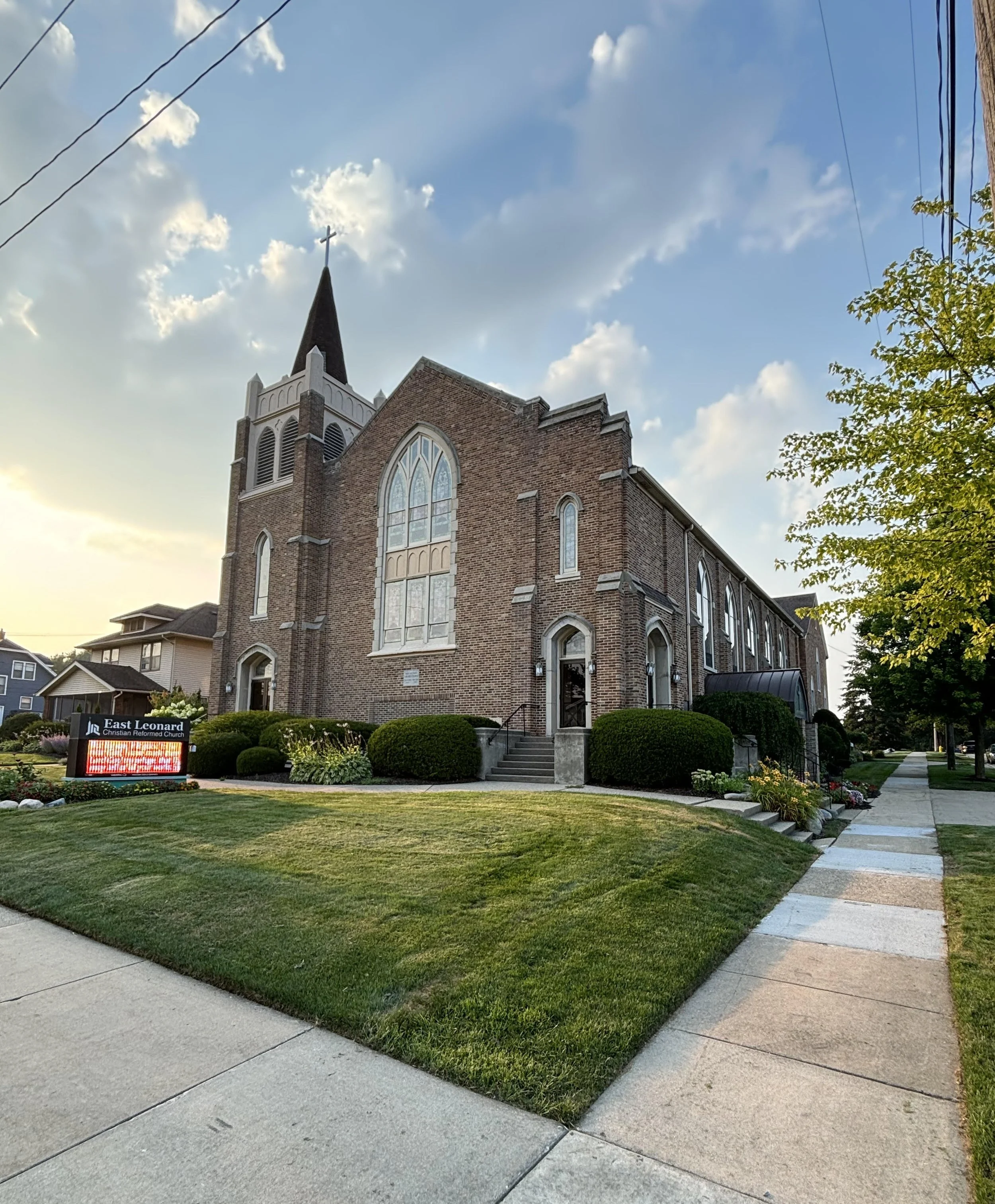 A brick church with a steeple topped with a cross, large arched stained glass windows, and a small set of stairs leading to the entrance. Surrounding the church are well-maintained lawns, bushes, and a sidewalk. A sign in front reads 'East Leonard Christian Reformed Church.' The sky is partly cloudy with sunlight illuminating the scene.