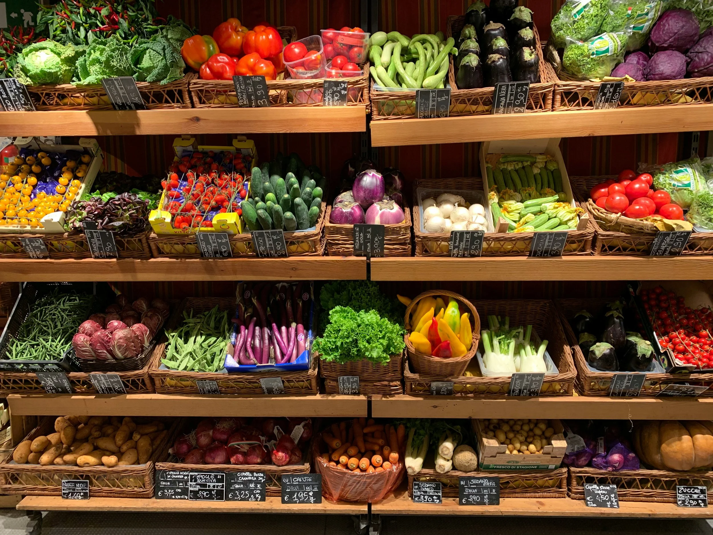 Display of various fresh vegetables arranged in baskets at a grocery store, including tomatoes, cucumbers, lettuce, purple eggplants, radicchio, onions, and carrots.