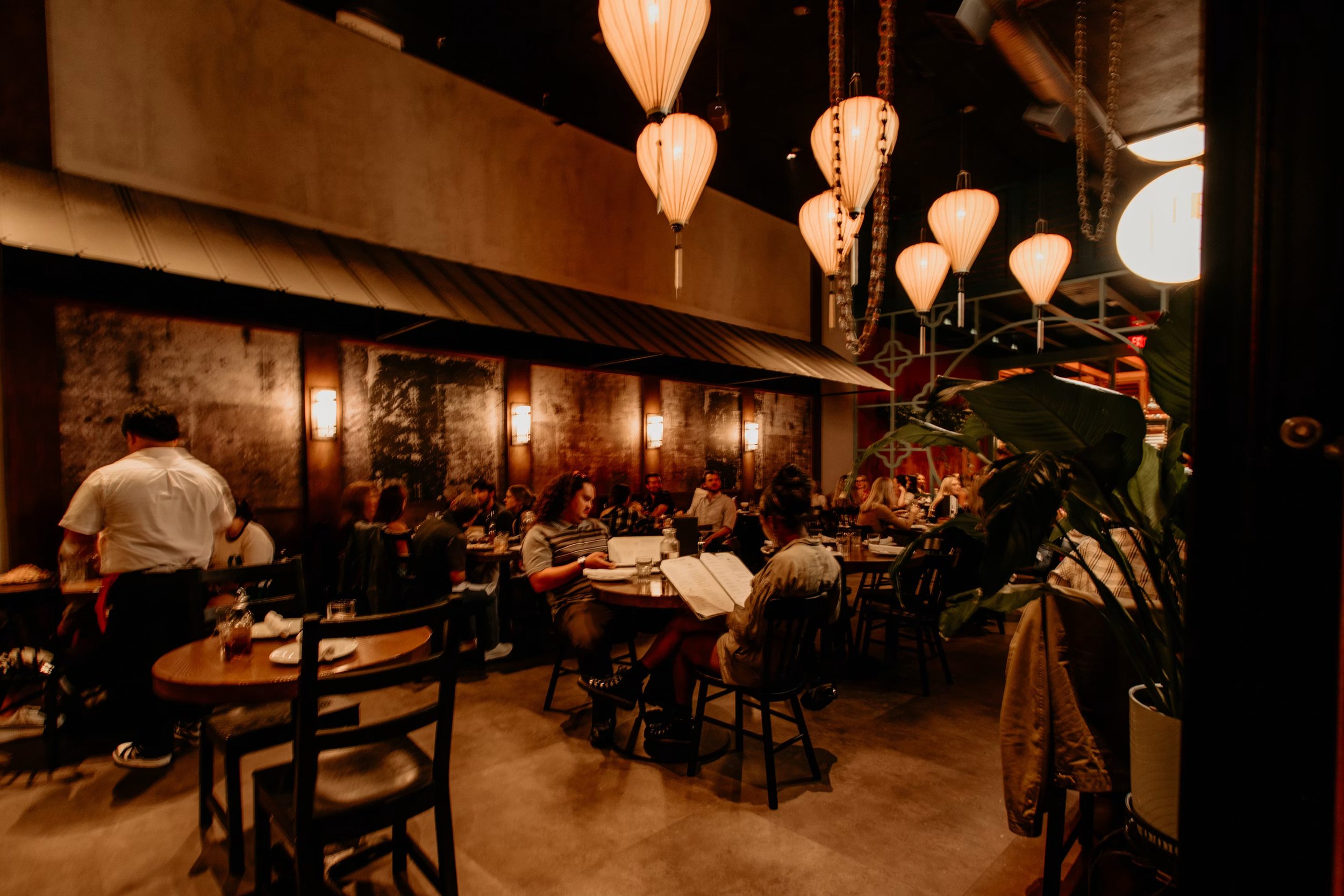 A crowded restaurant with warm lighting, hanging pendant lights, and people sitting at tables, dining and looking at menus.