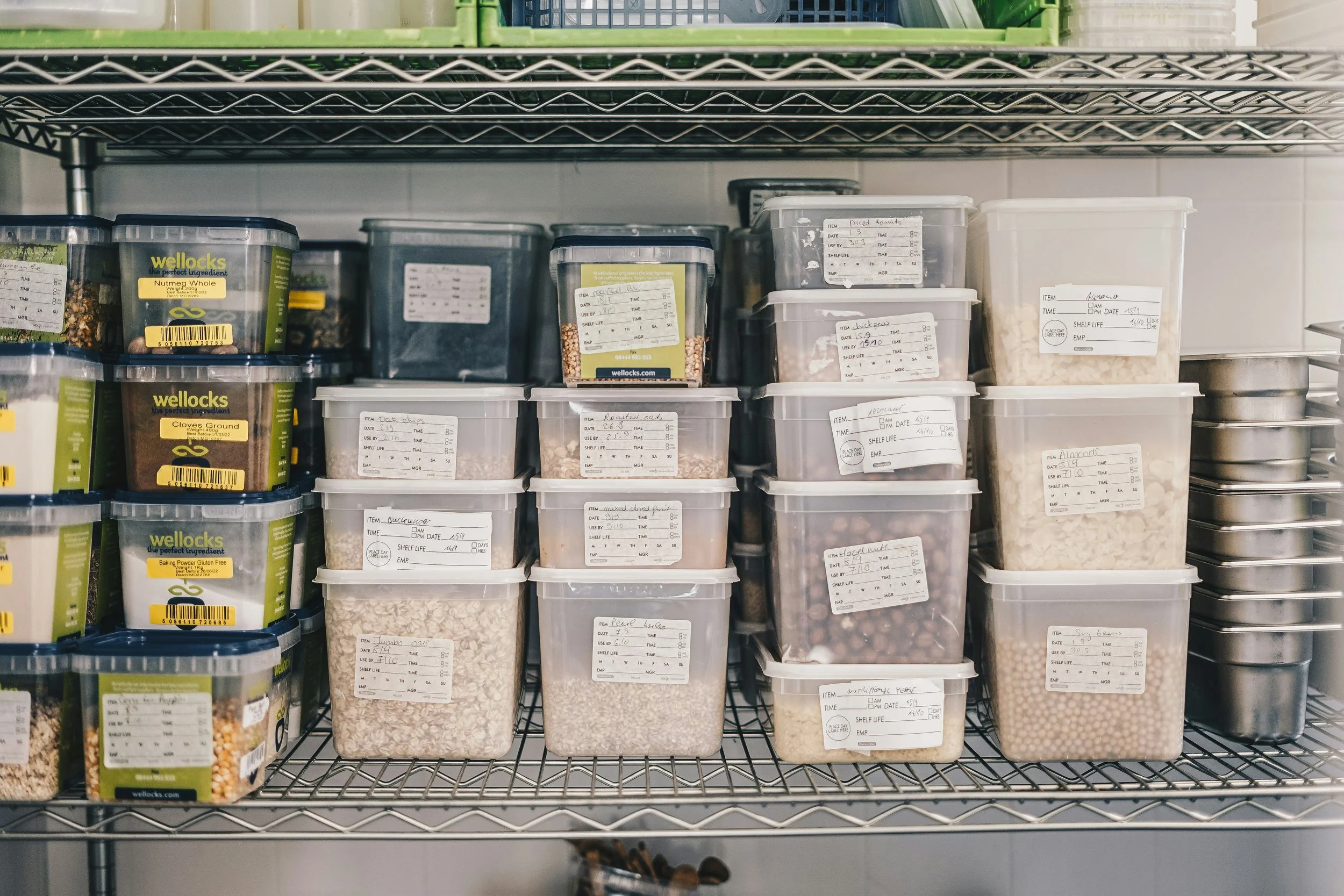 Clear plastic containers and jars filled with various dried foods and ingredients, labeled with handwritten notes, on metal shelving in a storage room.