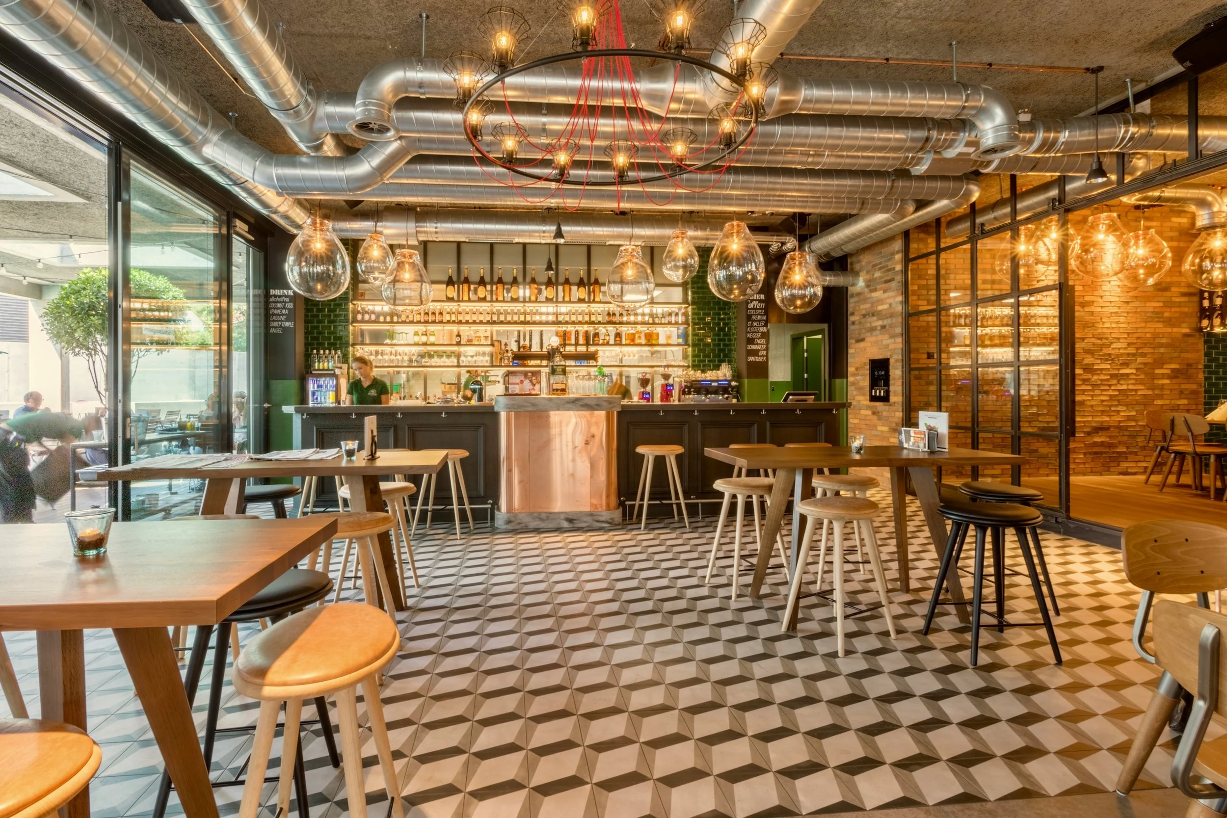 Interior of a modern café or restaurant with wooden tables and chairs, hanging light fixtures, patterned tile flooring, exposed ductwork and brick walls, and a bar area with bottles and glasses in the background.