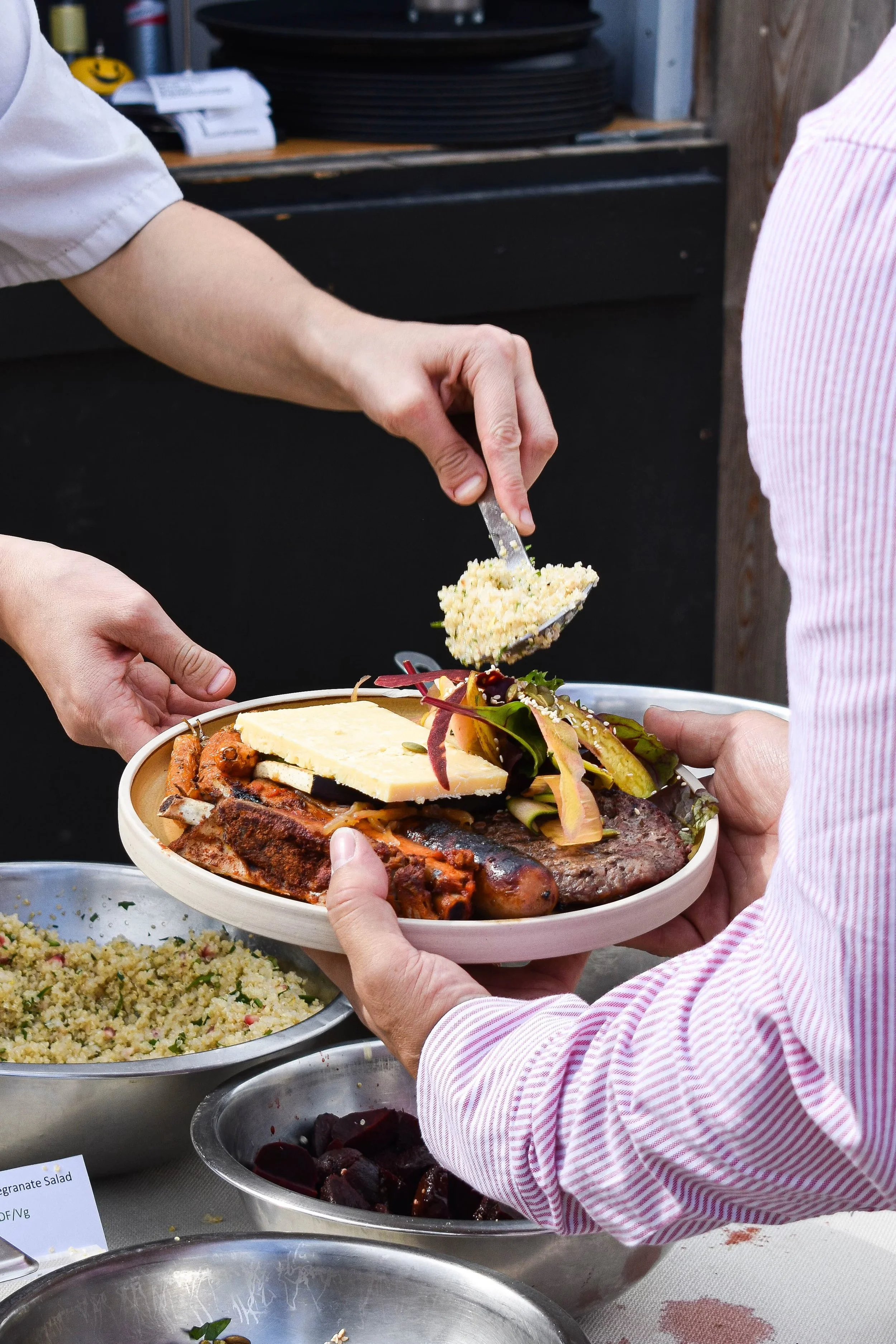 A person is holding a plate of food, including grilled meats, cheese, grilled vegetables, and salad. Another person is serving rice onto the plate. Various bowls of side dishes are visible on the table.