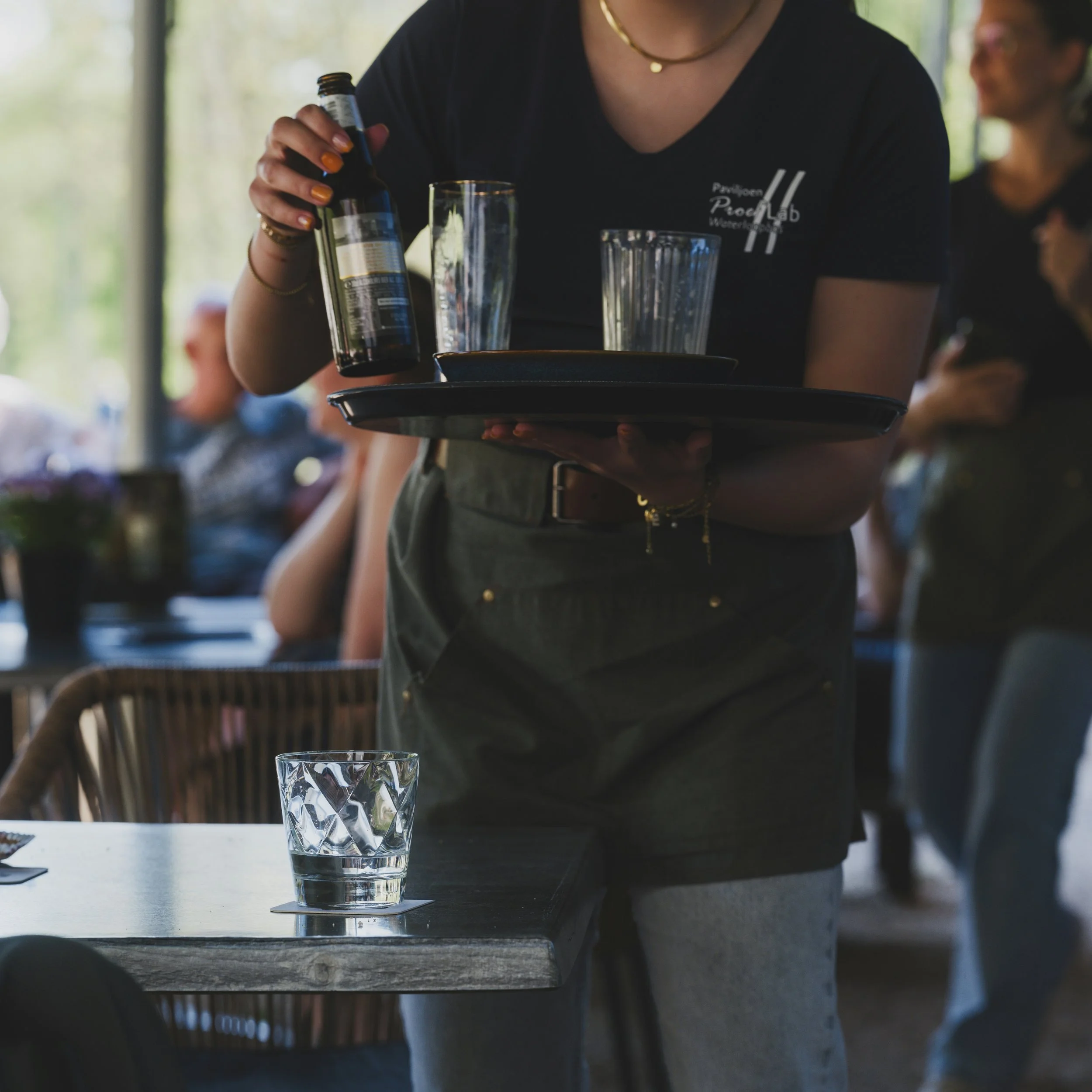 A person serving a drink from a small bottle into a tall glass on a table at a restaurant or café. The person is wearing a dark shirt and gold jewelry, and there are two other glasses on the table. The background is blurred with natural light coming through windows.