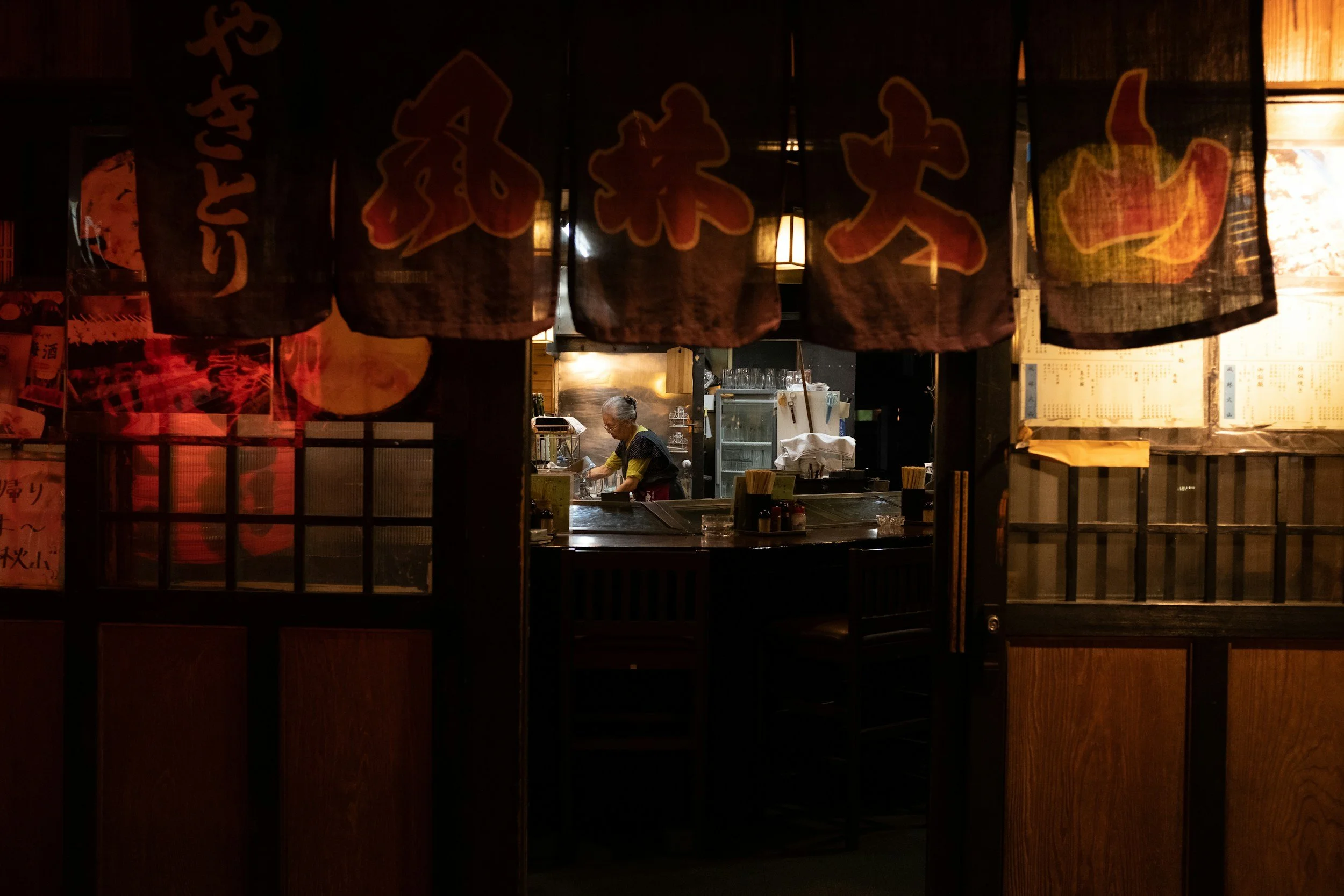A woman working behind the counter of a Japanese restaurant, visible through a dark curtain with Japanese characters, with warm lighting and wooden decor.