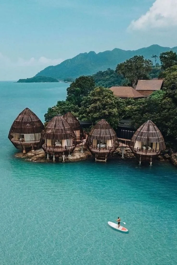 Overwater bungalows with thatched roofs on a tropical coastline, surrounded by lush greenery and mountains, with a person paddleboarding in turquoise water.