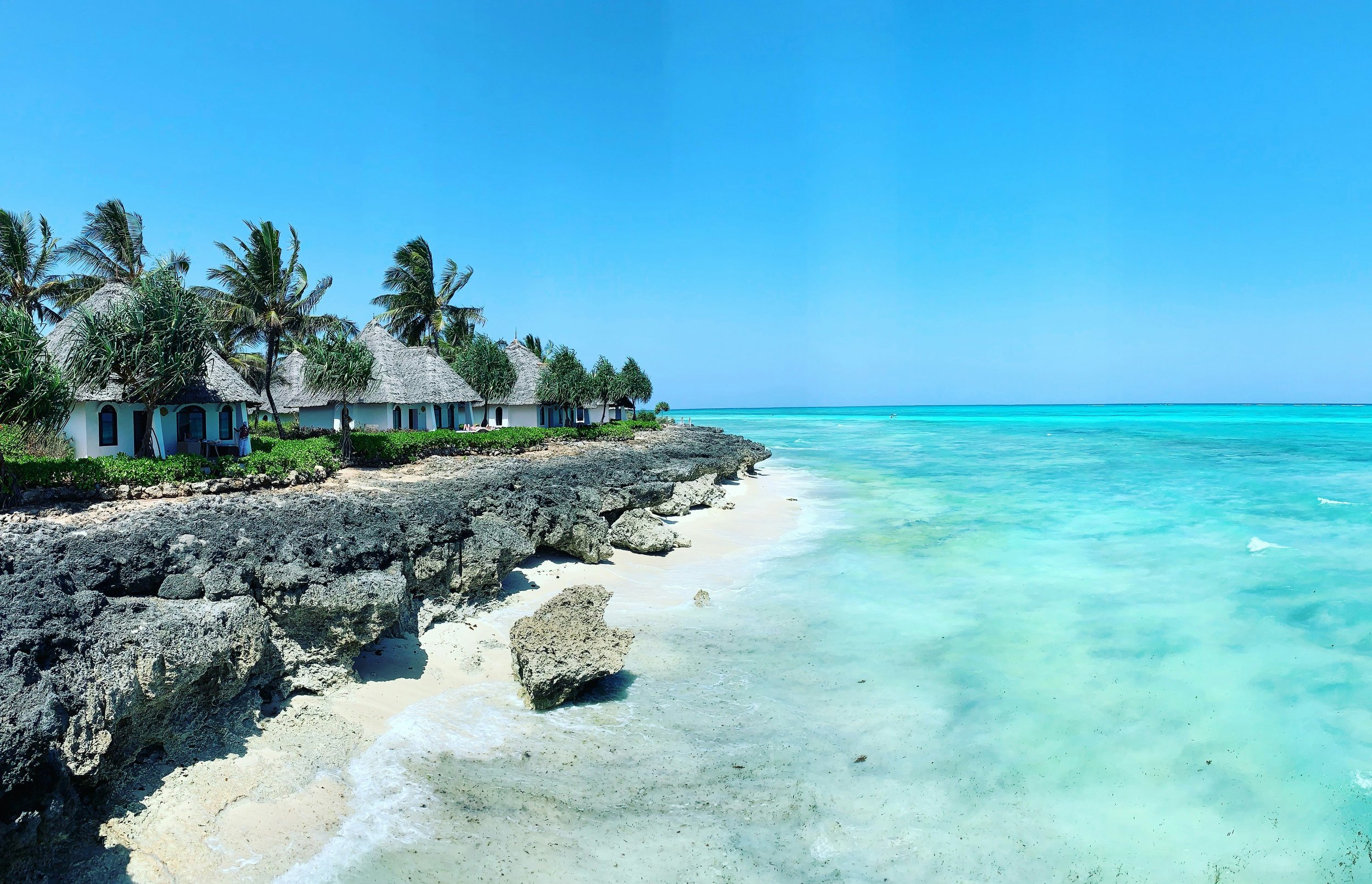 Beach in Tanzania with turquoise water, rocky shoreline, thatched cottages with palm trees, and a clear blue sky.