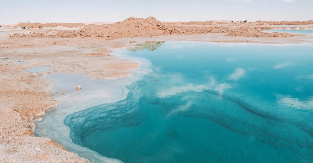A landscape in Siwa Oasis Egypt of a desert-like area with a large blue geothermal hot spring or pool, surrounded by reddish-brown terrain and distant hills.