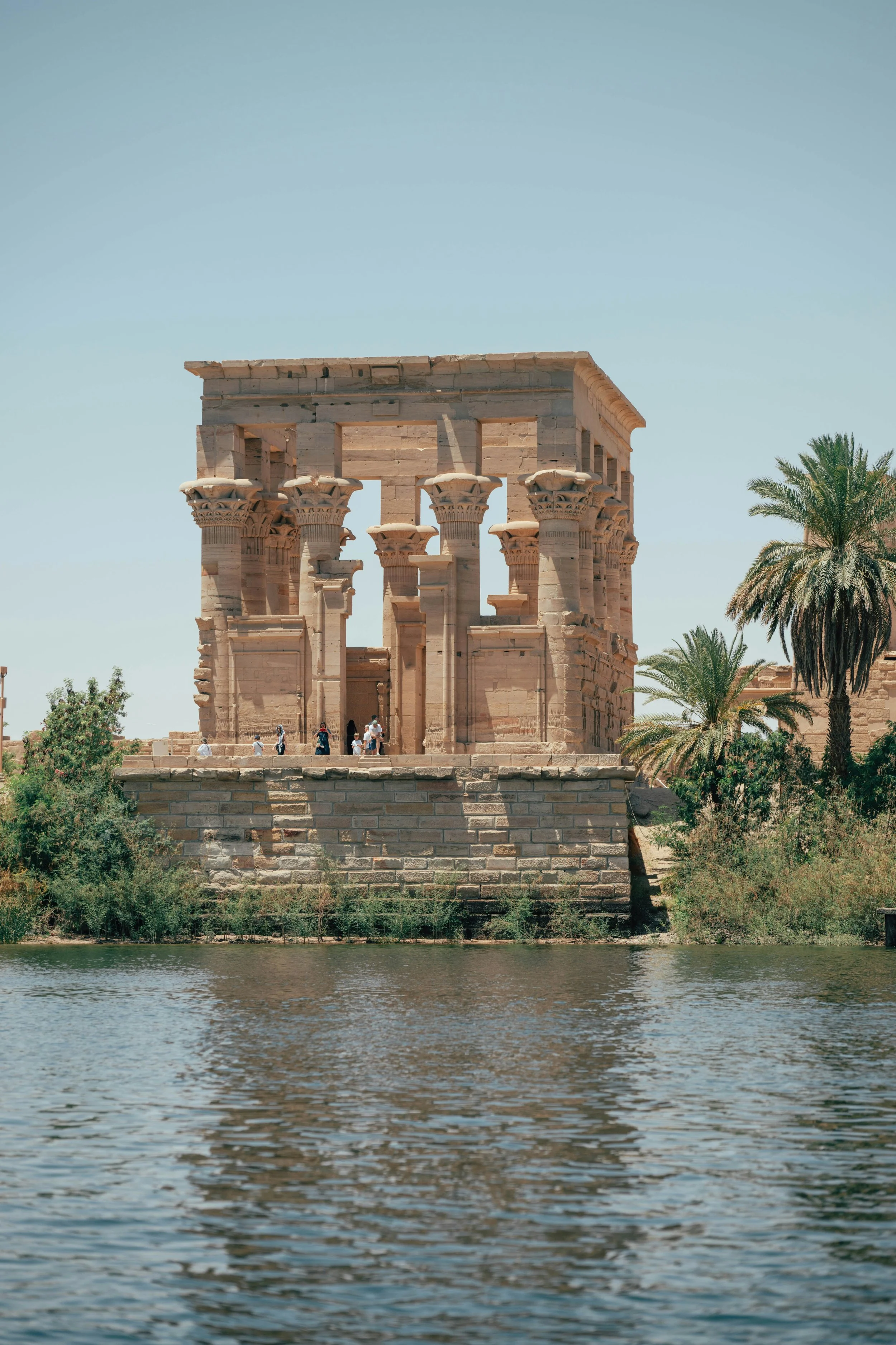 Ancient Egyptian temple ruins and palm trees along the banks of the Nile River under a clear sky.