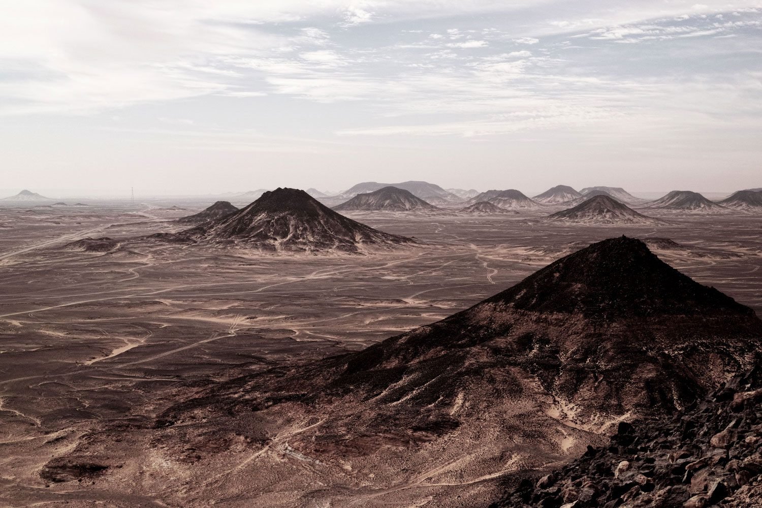 The black desert in Egypt with numerous dark, volcanic-looking mountains under a cloudy sky.