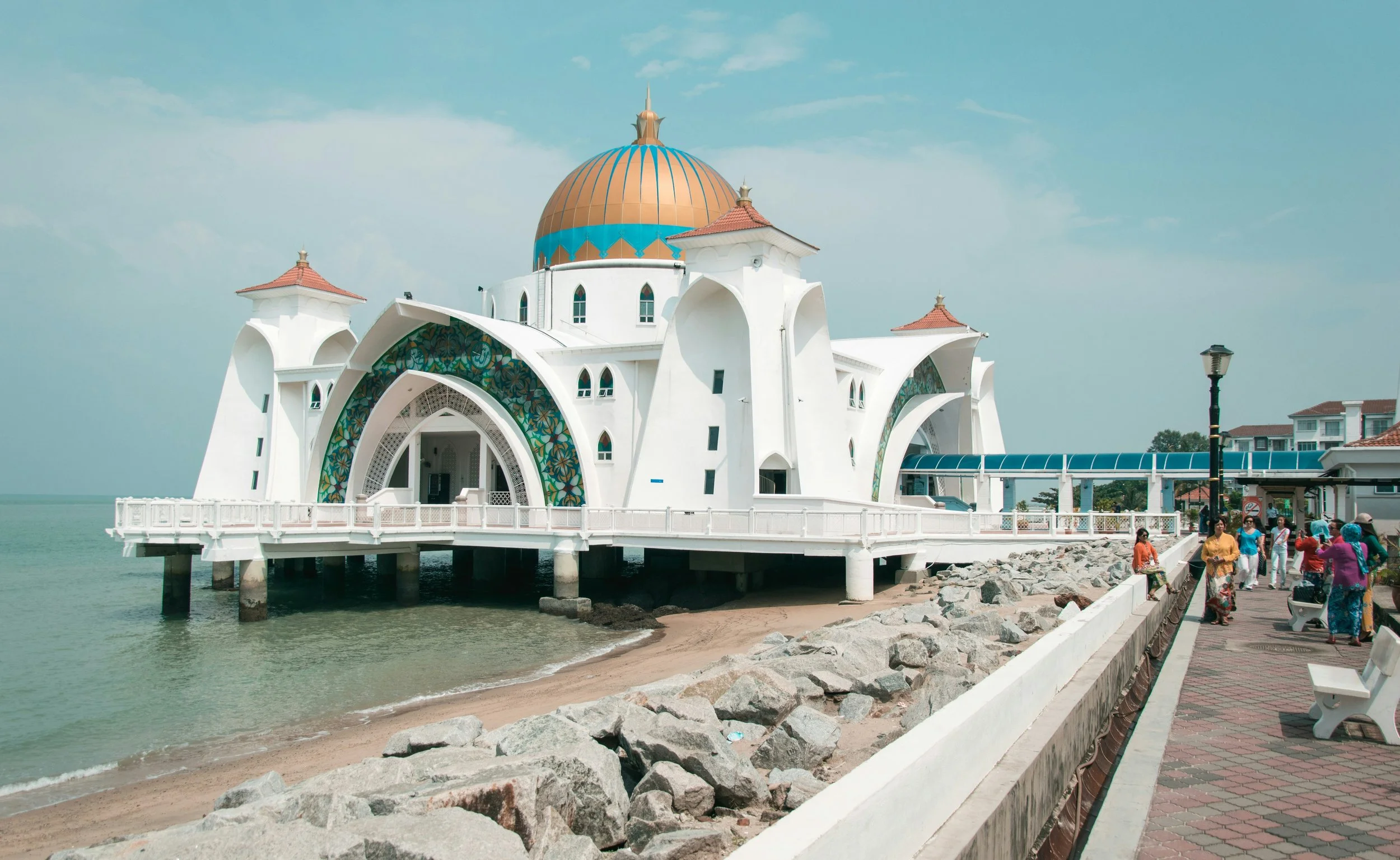 A Mosque in Malaysia with a large golden dome and smaller red-roofed turrets, built on stilts over water, with a walkway and people walking nearby.