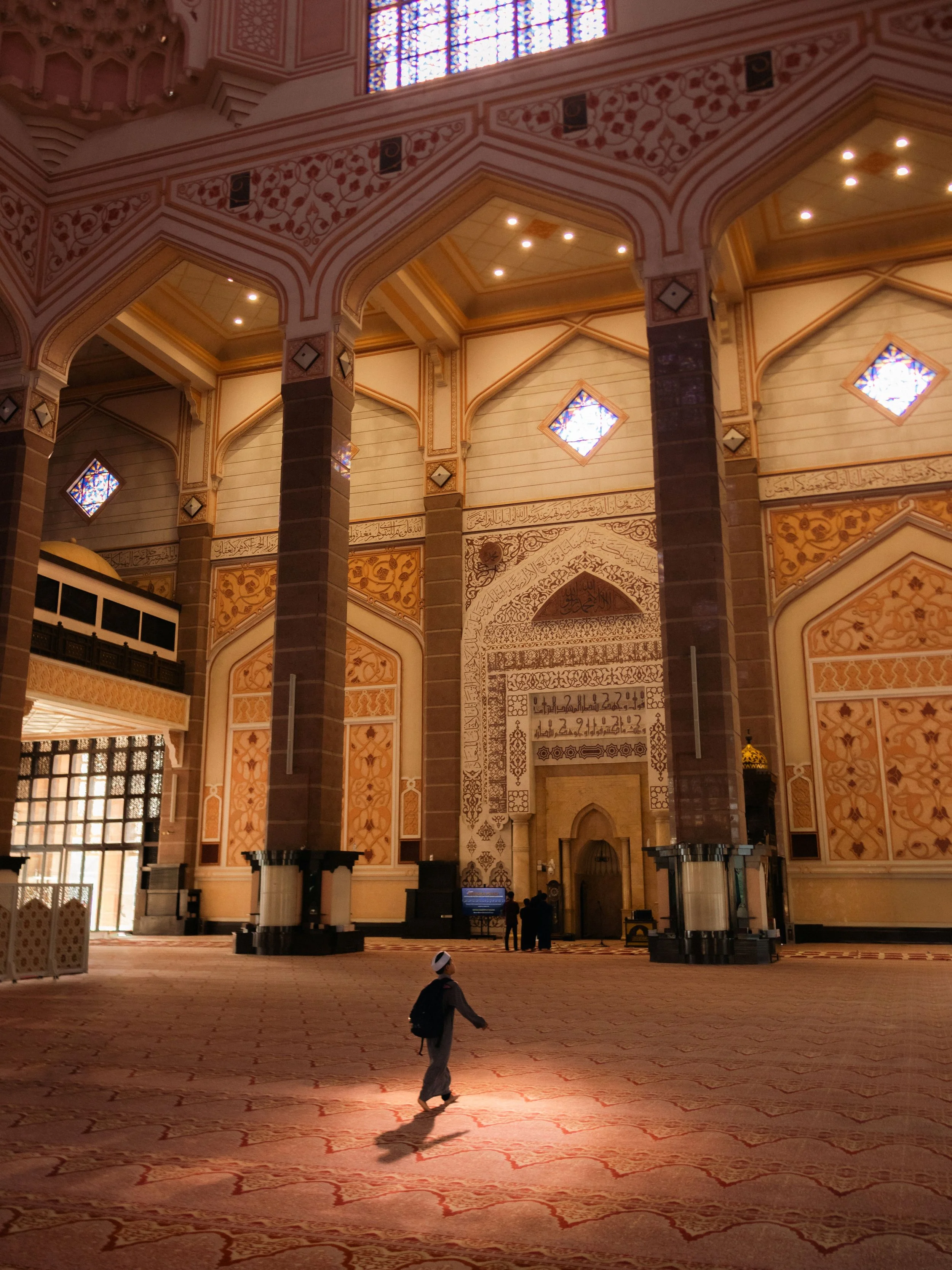 A white mosque on stilts over the ocean with a large copper-colored dome and smaller red-tiled roofs, surrounded by a paved walkway with people walking and gathering, some wearing colorful clothing.