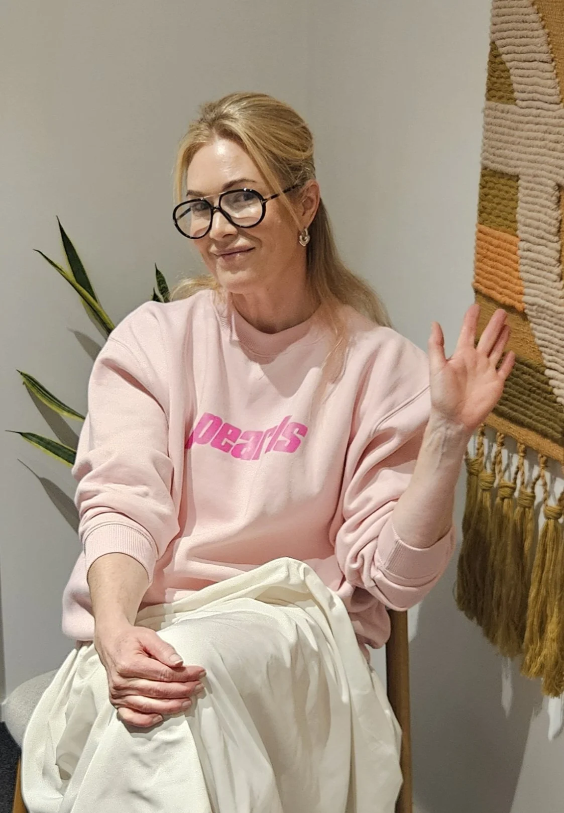 A woman with blonde hair, glasses, and earrings sitting on a chair in front of a light-colored wall. She is wearing a pink sweatshirt with red text and white pants, smiling, and gesturing with her right hand. There is a green plant behind her and a decorative wall hanging on her right.