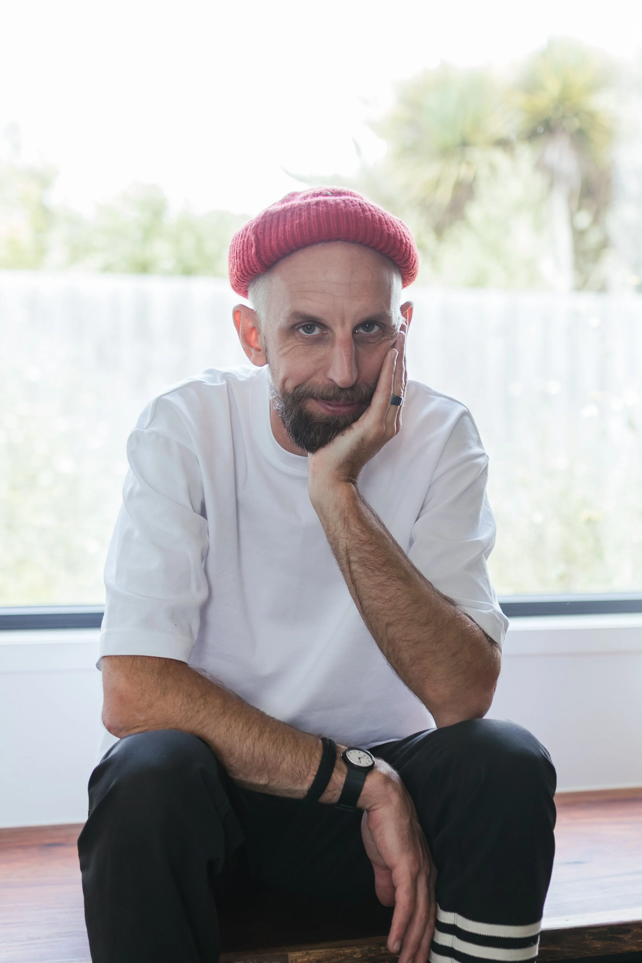 A man with a red knit beanie, beard, and mustache sitting by a large window, resting his head on his hand, wearing a white t-shirt, watch, and black pants.