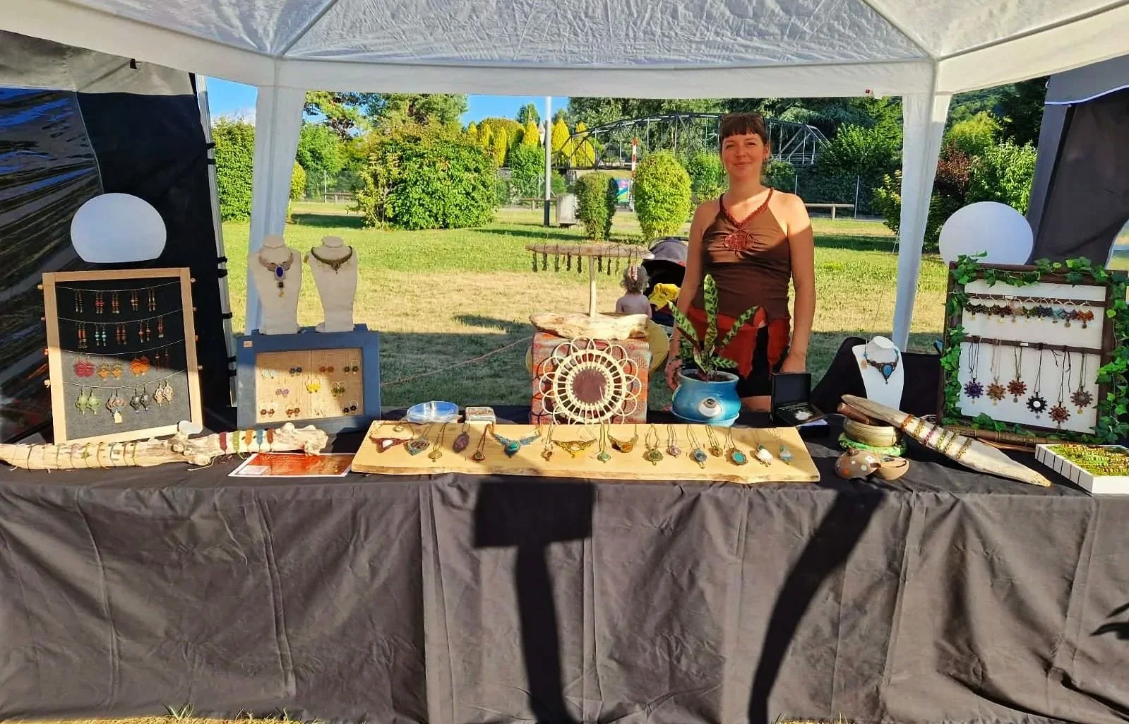Une femme se tient derrière un stand de bijoux fait main présenté en extérieur, avec plusieurs colliers, bracelets et boucles d'oreilles en vitrine et exposés sur la table.