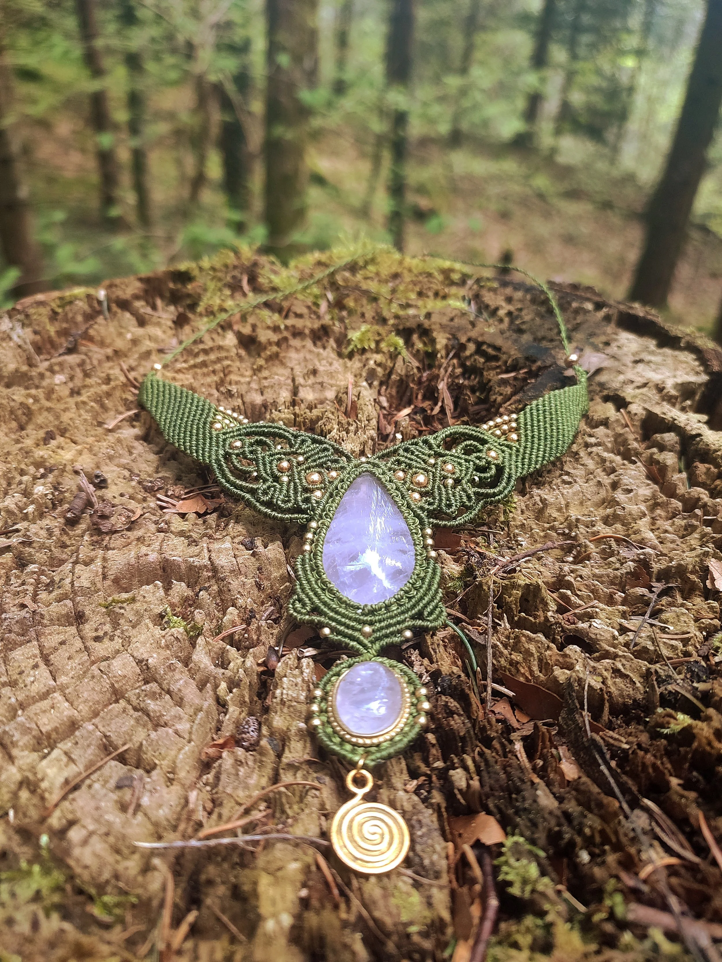 Un collier fait main avec un pendentif en forme de larme en pierre de lune, entouré de filigranes en fil vert et or, posé sur une branche dans une forêt