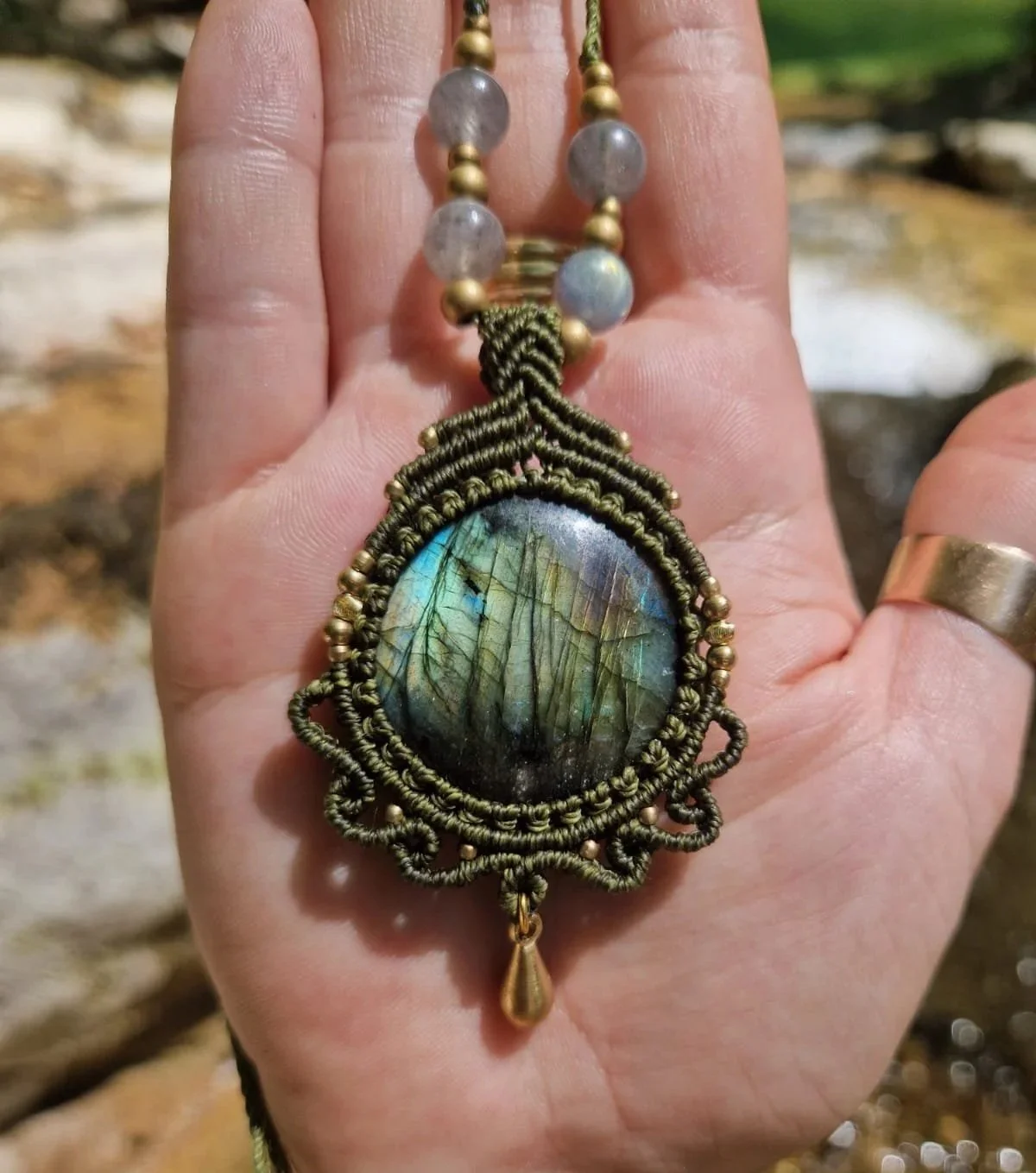 Un pendentif avec une pierre en labradorite entourée de fil de macramé brillant, porté sur une main avec une bague en or.