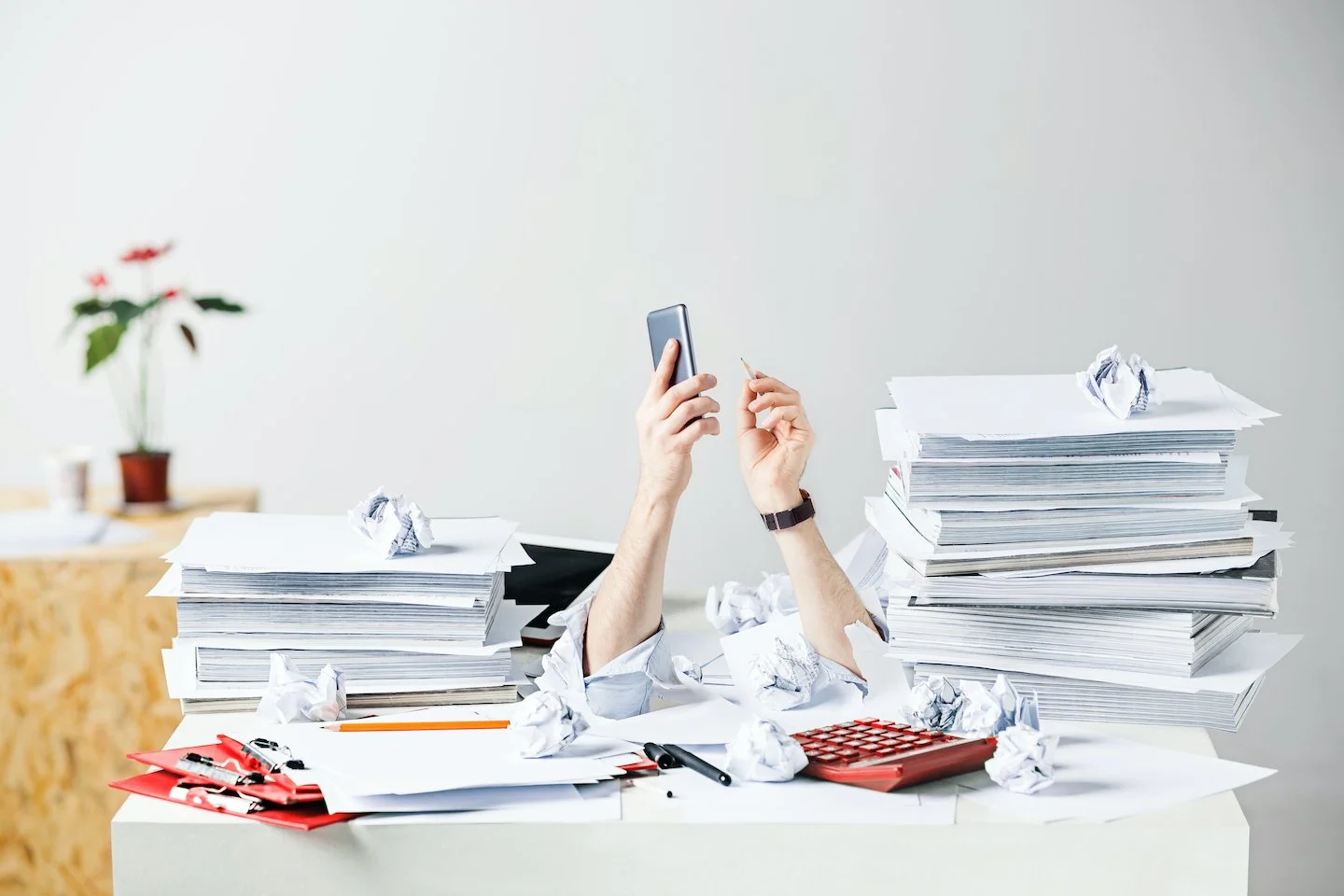 Two hands reaching up from a very messy desk stacked with two very high piles of paper.