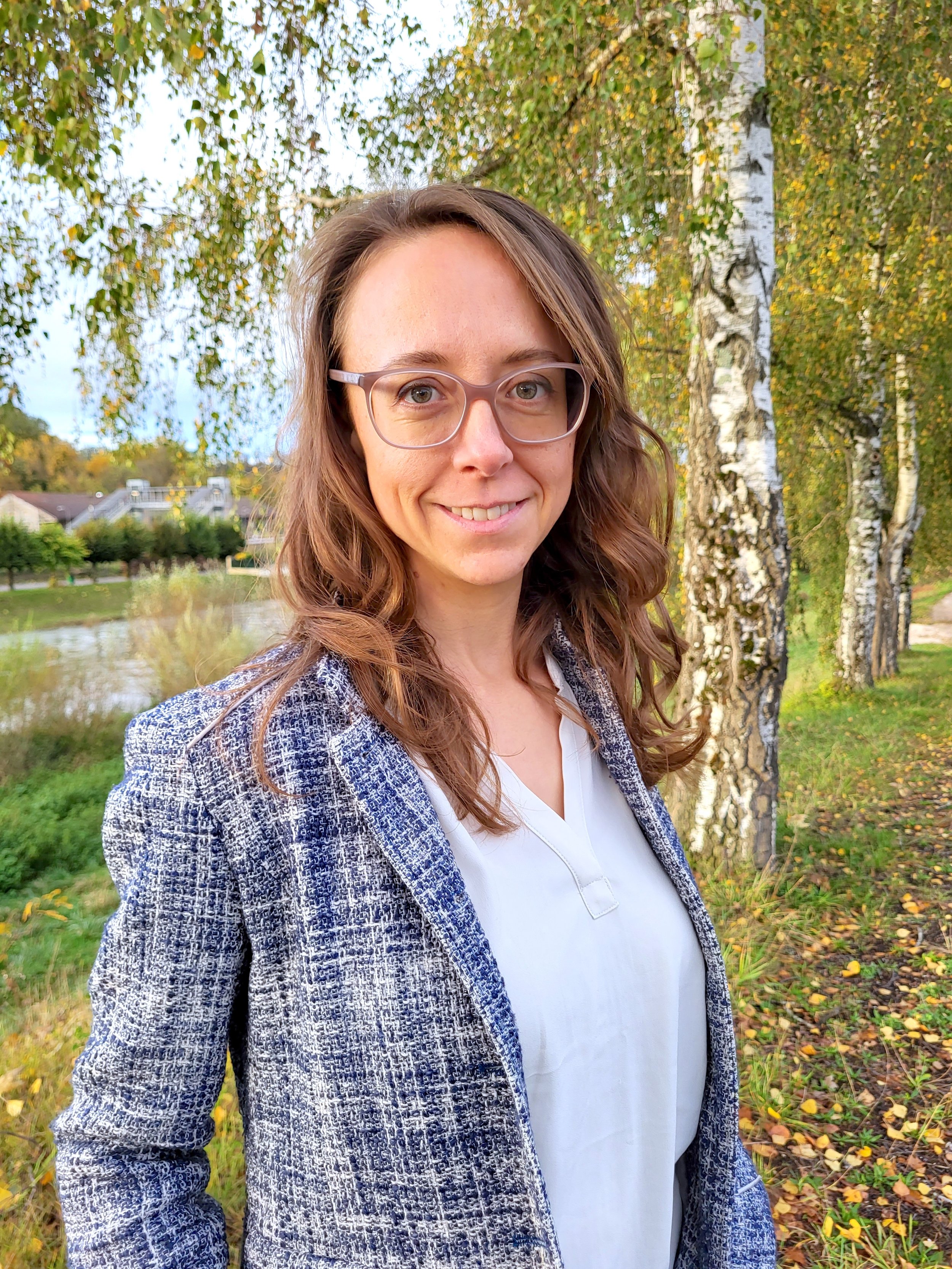 Tammy Mayer is standing outside with birch trees and a river behind her. There are yellow leaves on the ground. She is wearing a blue and white blazer with a white shirt. She is wearing glasses and has a soft smile.