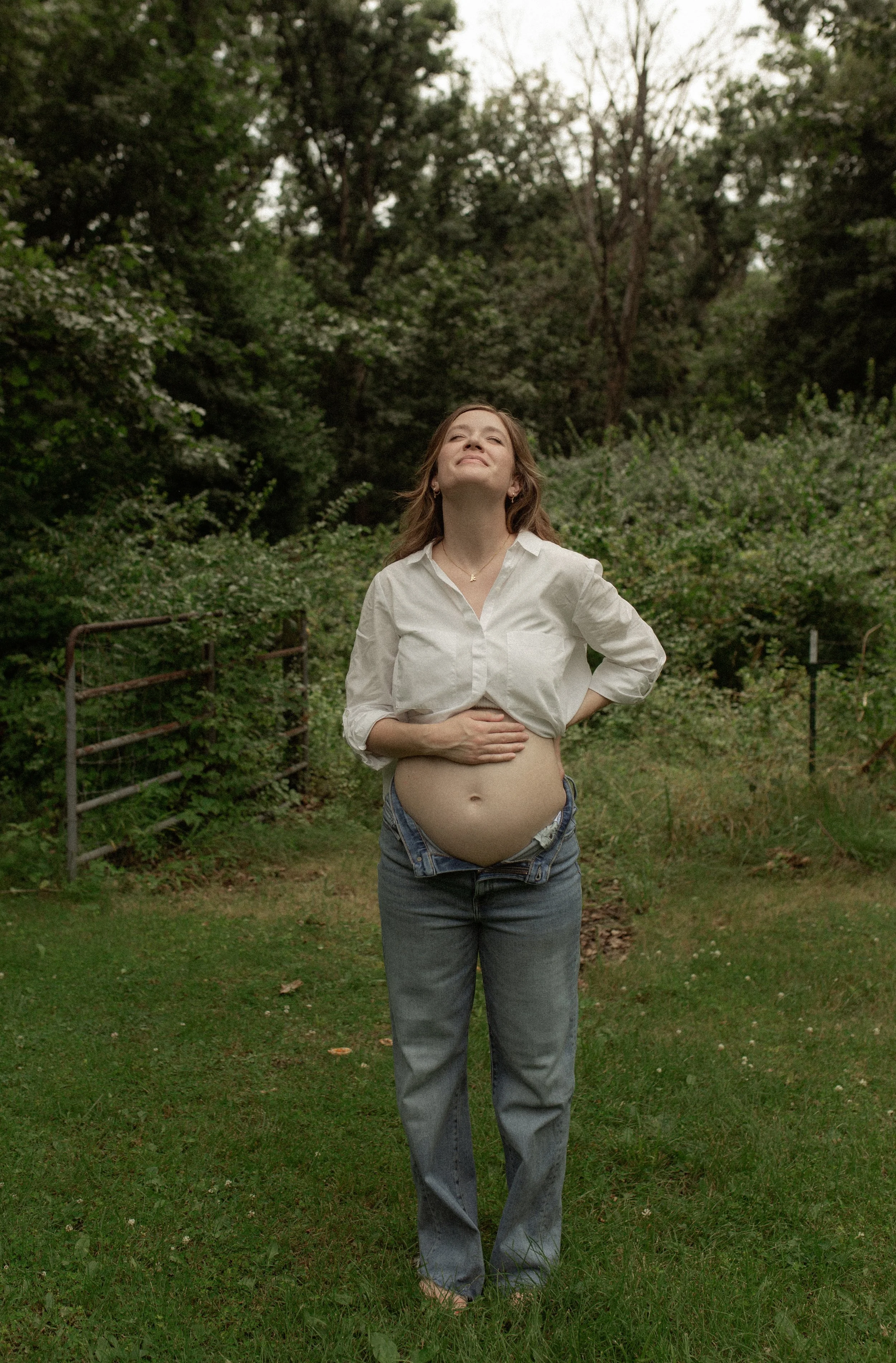 A woman stands outdoors in a green, wooded area, smiling with eyes closed, wearing a white shirt and blue jeans, exposing her pregnant belly with her hands resting on her stomach.
