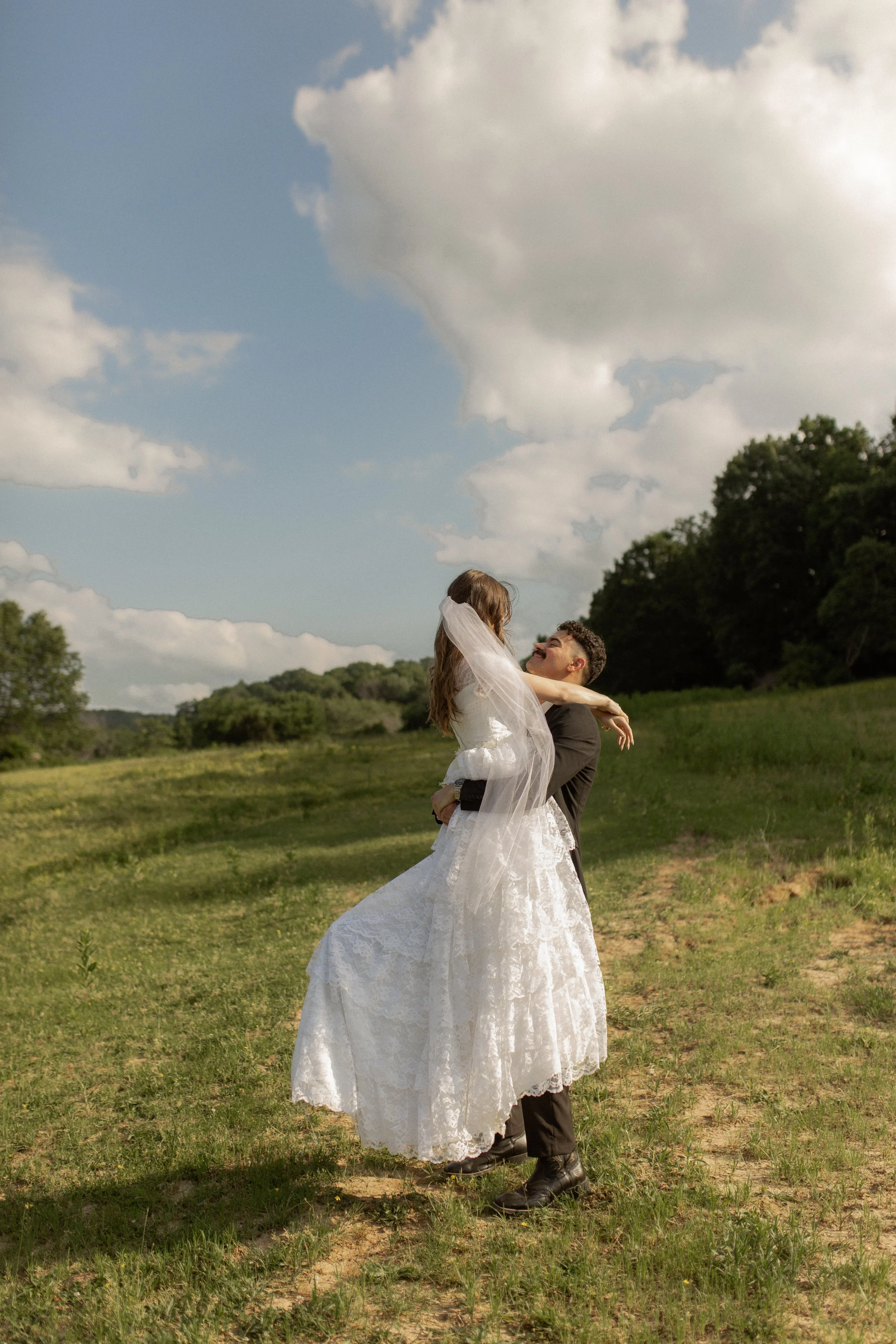 A bride and groom outdoor wedding photo, with the bride in a white wedding dress and veil, and the groom in a dark suit, standing on a grassy field with trees and partly cloudy sky in the background. The groom is lifting the bride, and they are smiling at each other.
