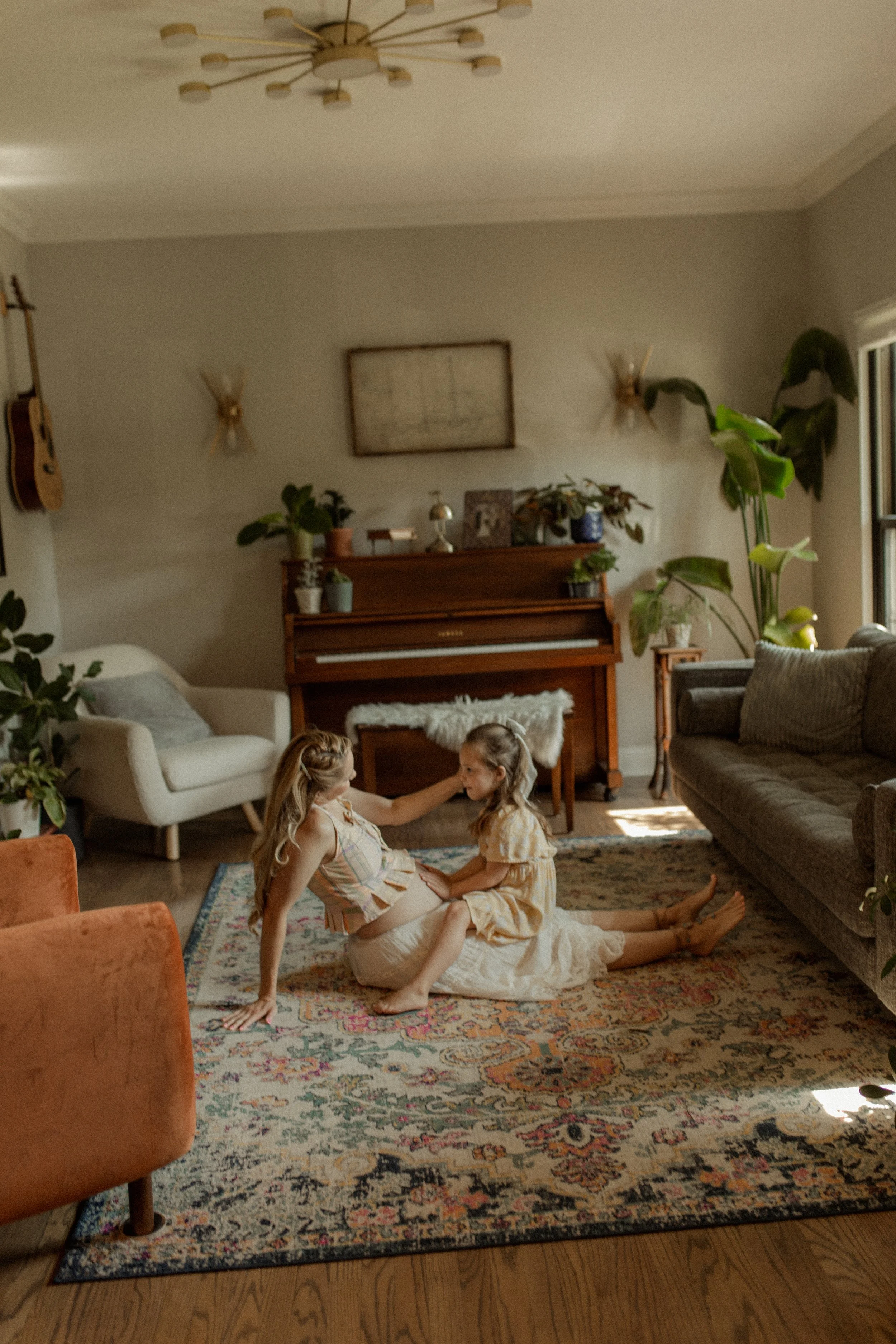 A living room with two young girls playing on a patterned rug. The room features a white sofa, a brown wooden piano with plants on top, a gray armchair, and various wall decorations including a framed picture and wall sconces. Sunlight is coming through a window on the right.