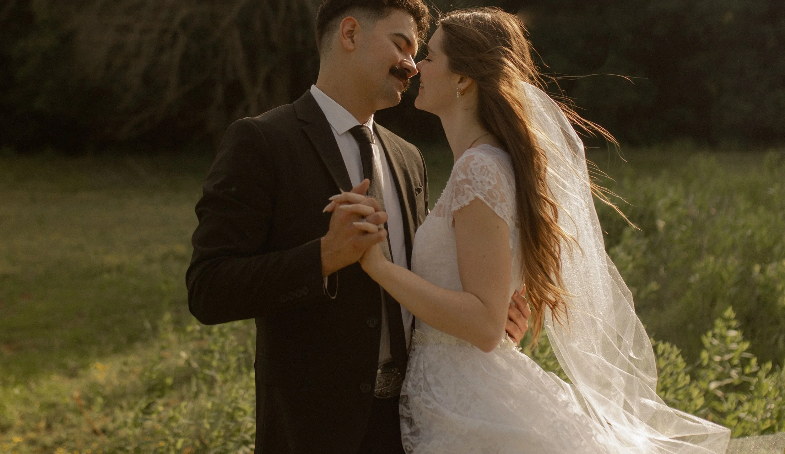 A bride and groom happily dance together outdoors during their wedding, holding hands and smiling with their foreheads touching, at sunset in a natural setting.