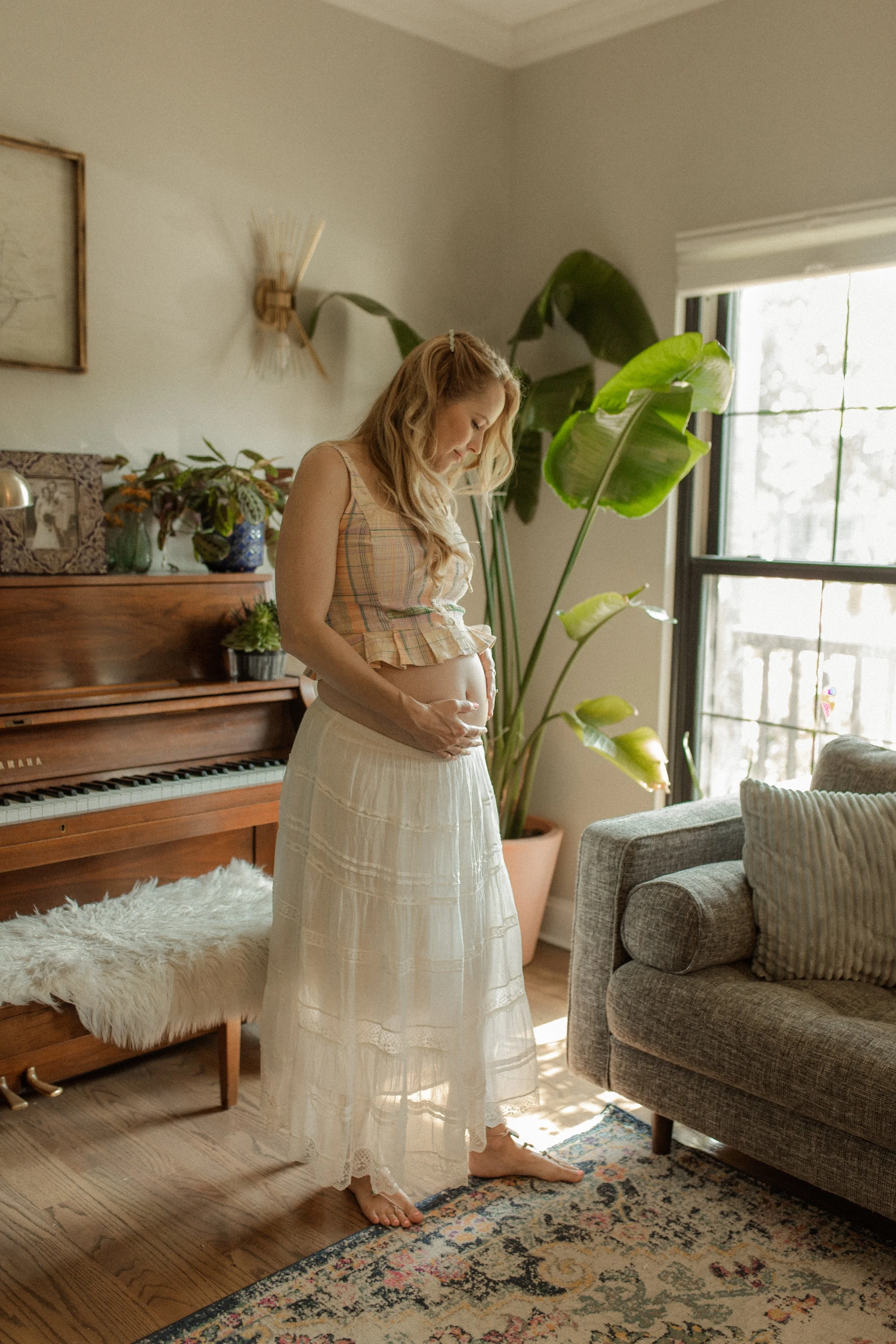 A pregnant woman stands barefoot in a cozy living room, gently touching her belly. She is wearing a sleeveless plaid top and a long white skirt. The room is filled with natural light from a large window, decorated with plants, a wooden piano decorated with plants, and a colorful rug on dark wood flooring.