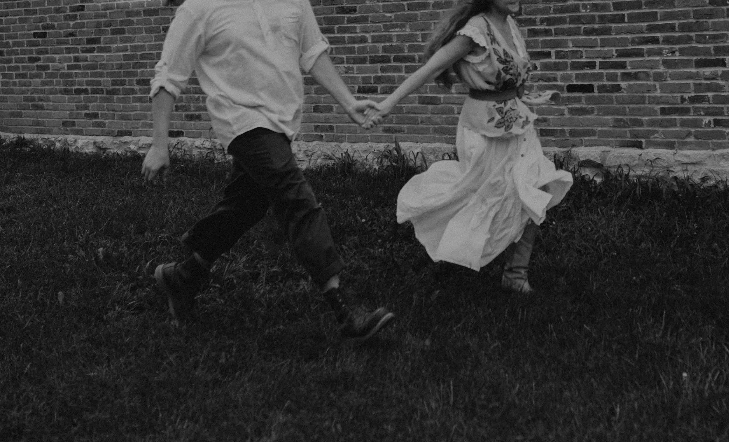 Two children holding hands and running outside with a brick wall in the background.