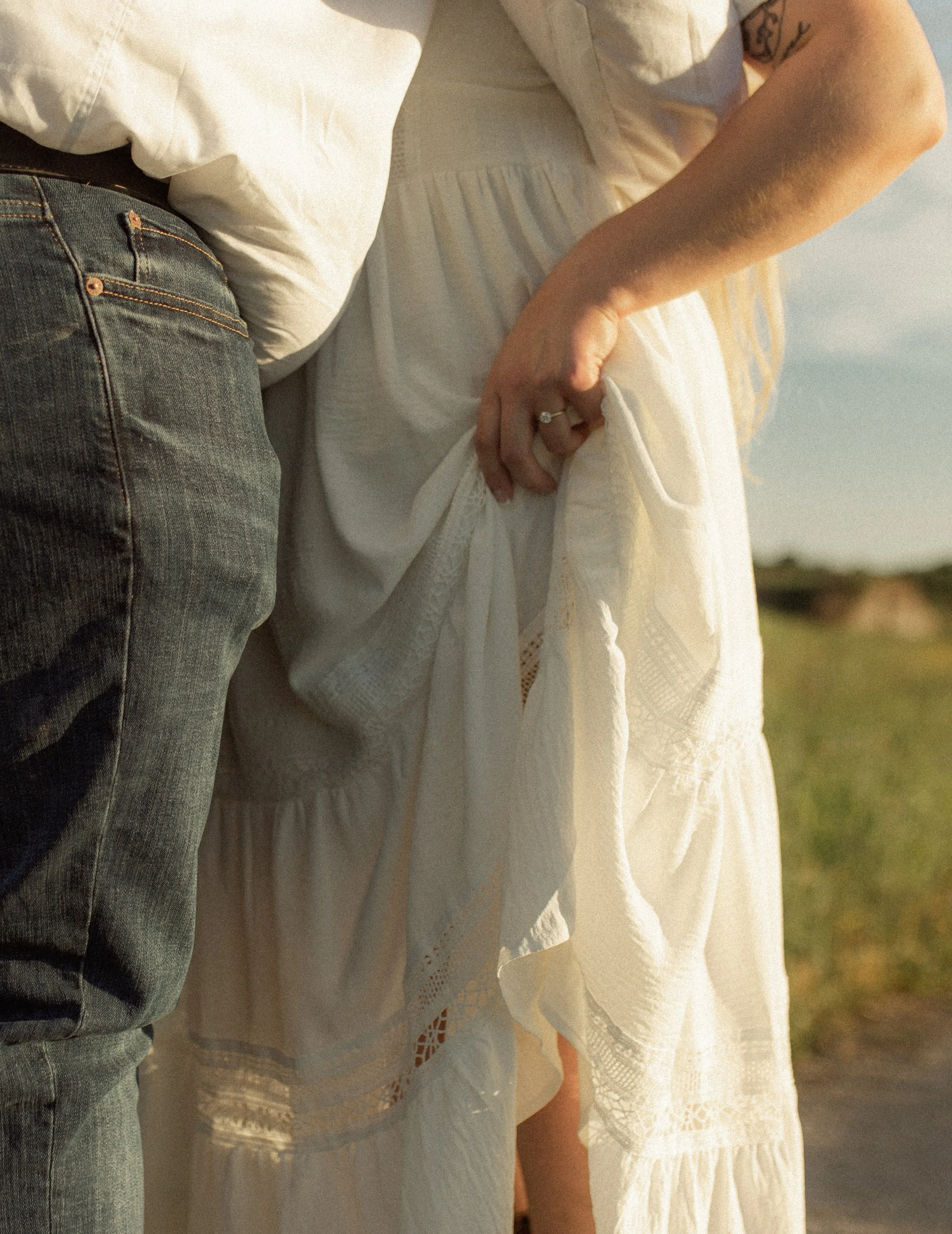 Close-up of two people holding hands, one wearing a diamond ring, with a white dress and a tattoo visible on one person's arm, against a sunset background.