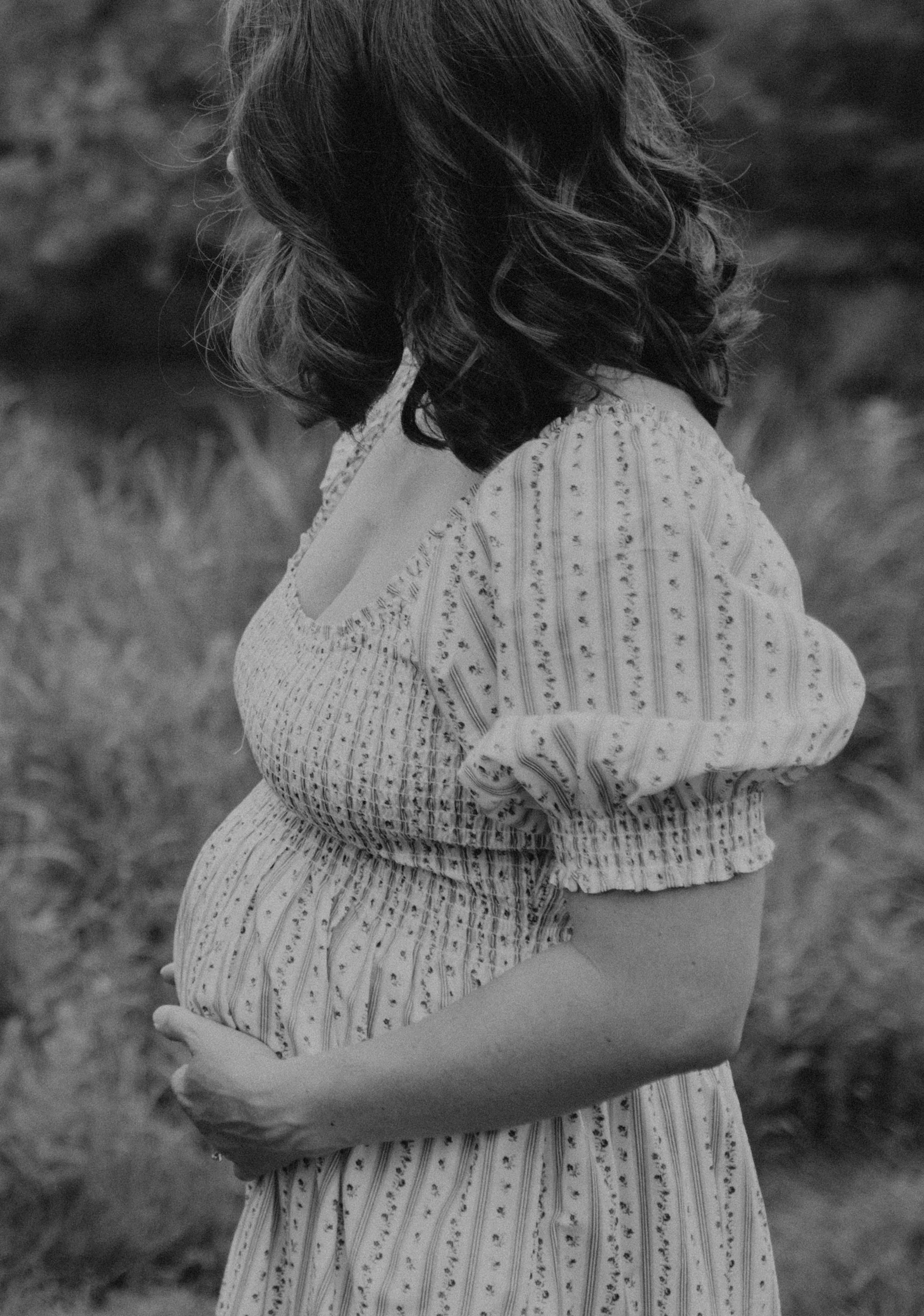 A black-and-white photo of a pregnant woman standing outdoors, cradling her belly with one hand, wearing a patterned, long-sleeved dress. Her face is not visible as the photo is taken from the back and side.