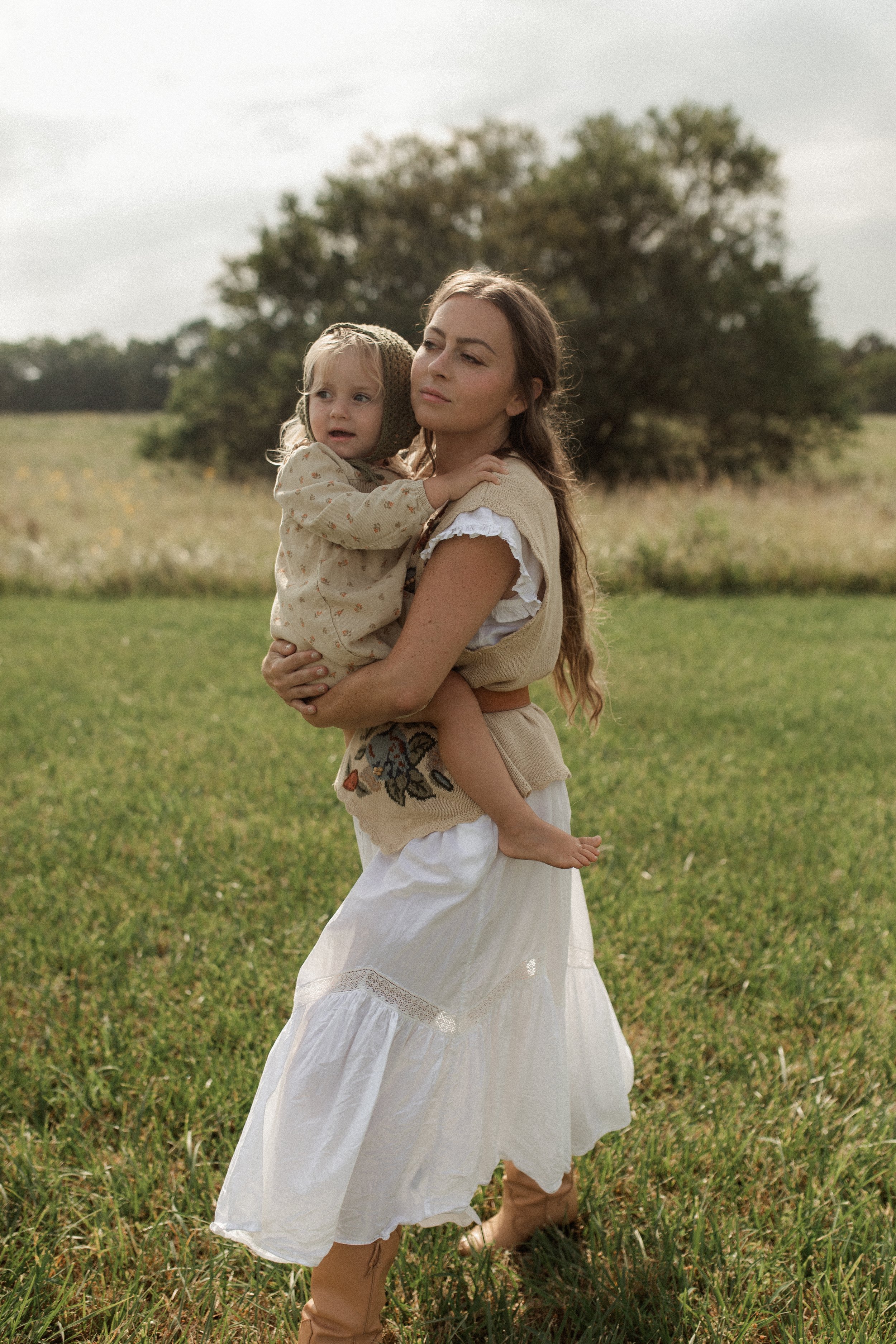 A woman wearing a white flowy skirt and a beige vest holding a young girl with blonde hair and a beige bonnet in an open grassy field with trees in the background.