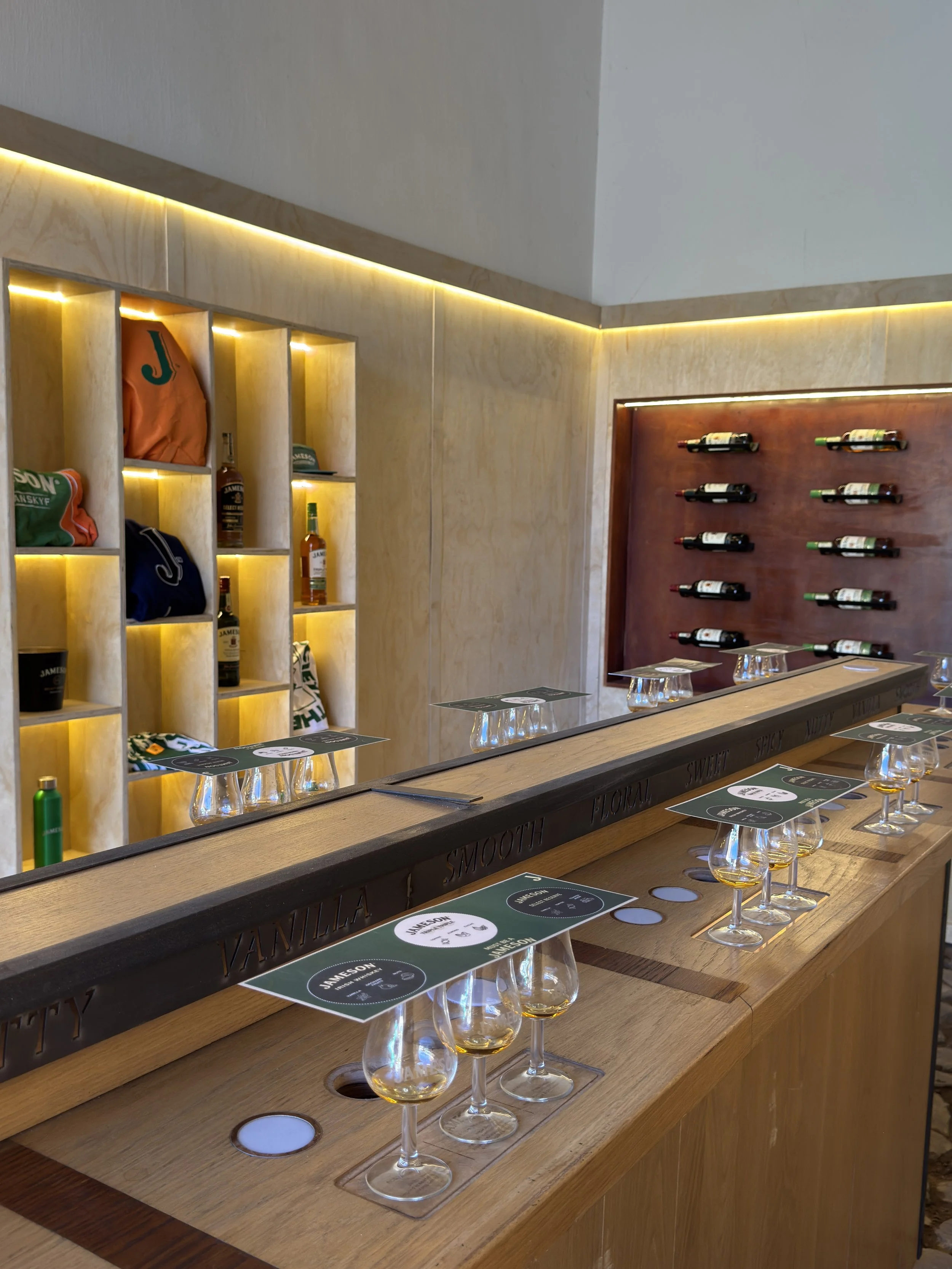 A wooden bar counter with wine bottles on a wall-mounted wine rack, glasses with whiskey, and paper menus.