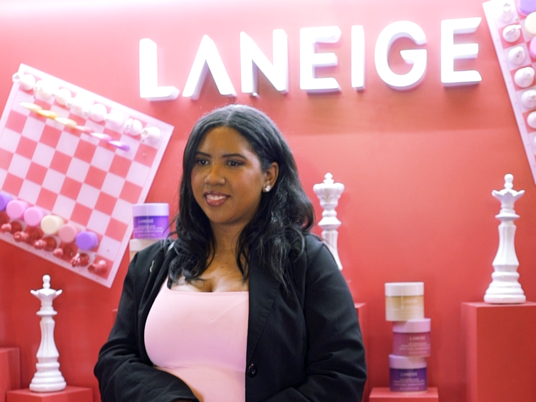 A woman with dark hair and a black blazer standing in front of a pink background with the word 'LANEIGE' and decorative chess piece props.
