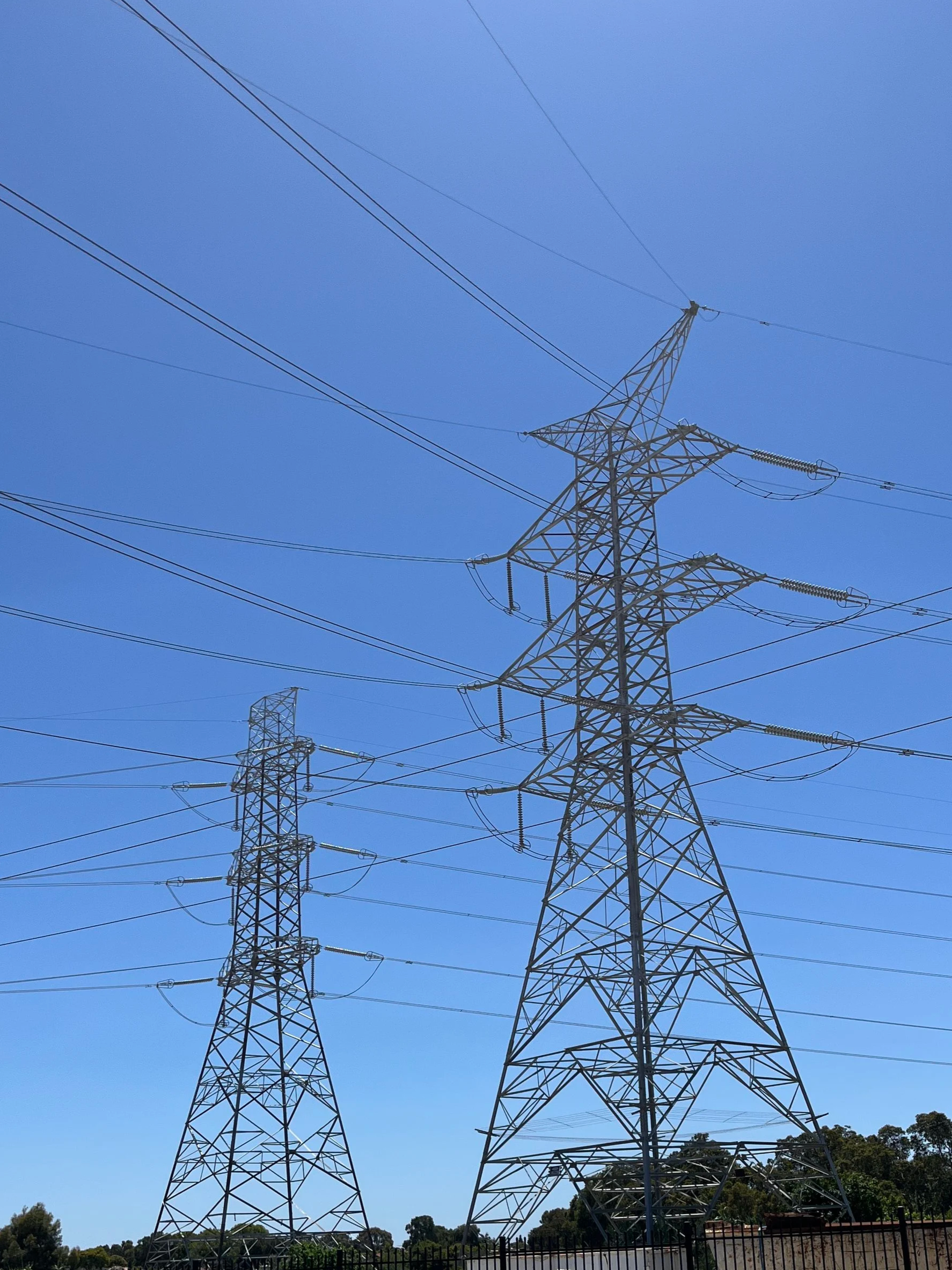 Several electrical transmission towers with power lines against a blue sky and a tree line in the background.