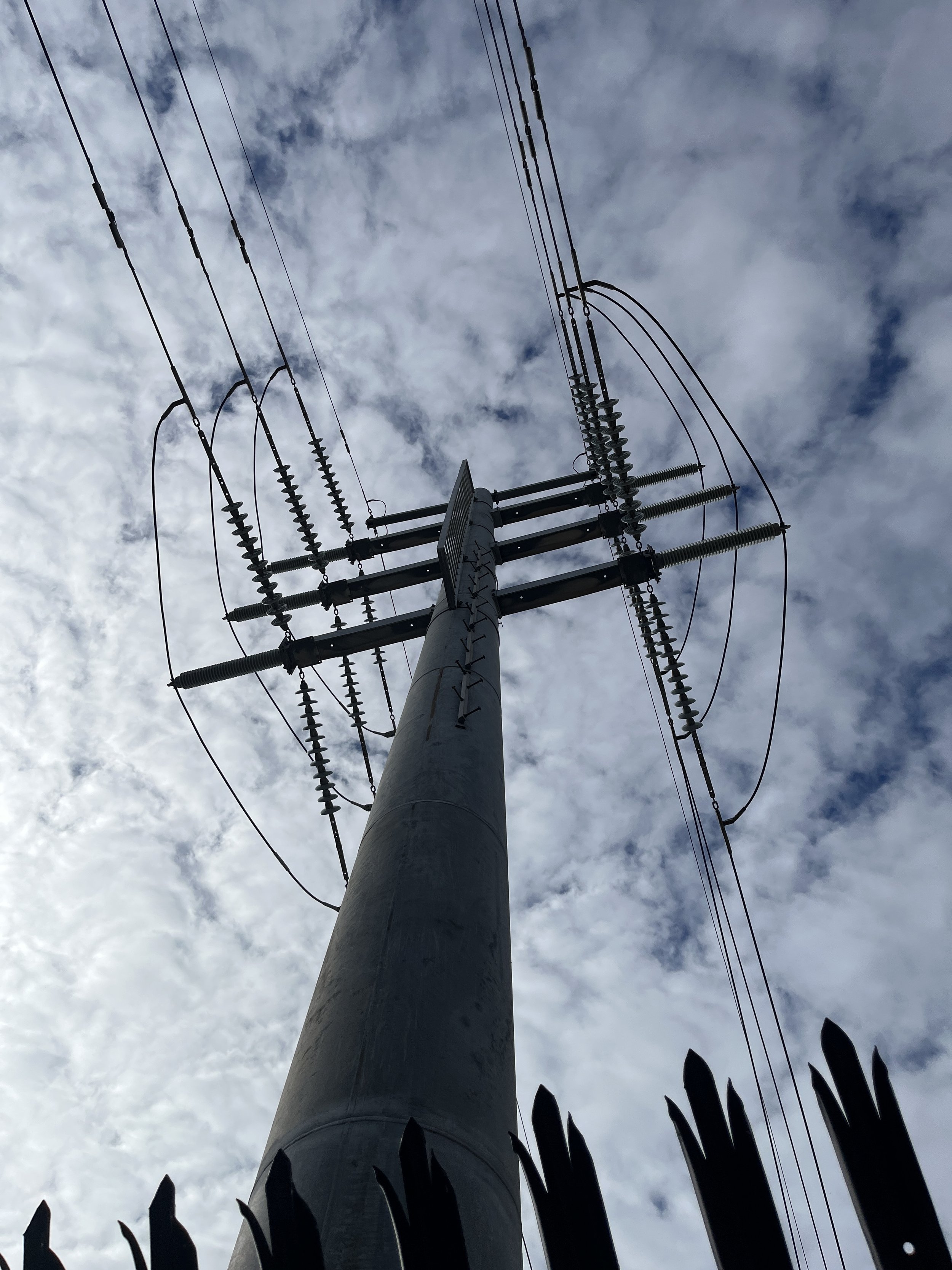 A tall utility pole with multiple electrical wires and insulators against a cloudy sky, with silhouettes of sharp-edged fence in the foreground.