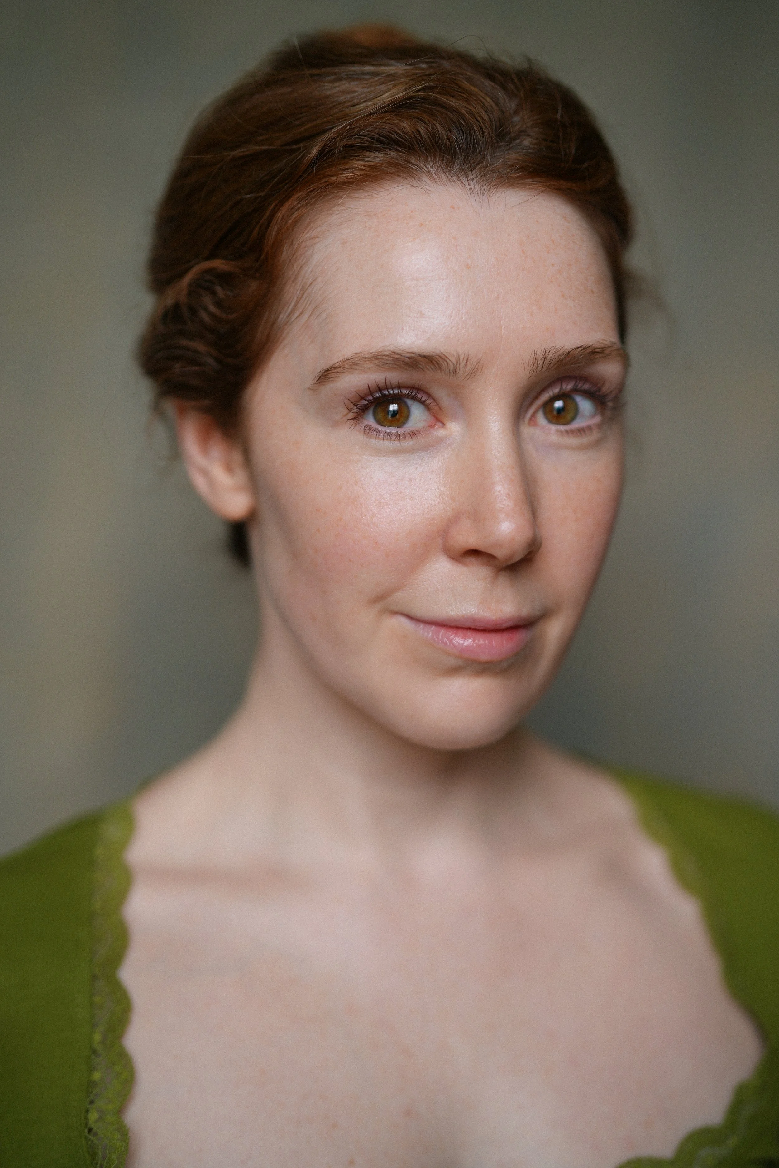 Close-up of a young woman with fair skin, red hair, and brown eyes, wearing a green top, standing against a neutral background.
