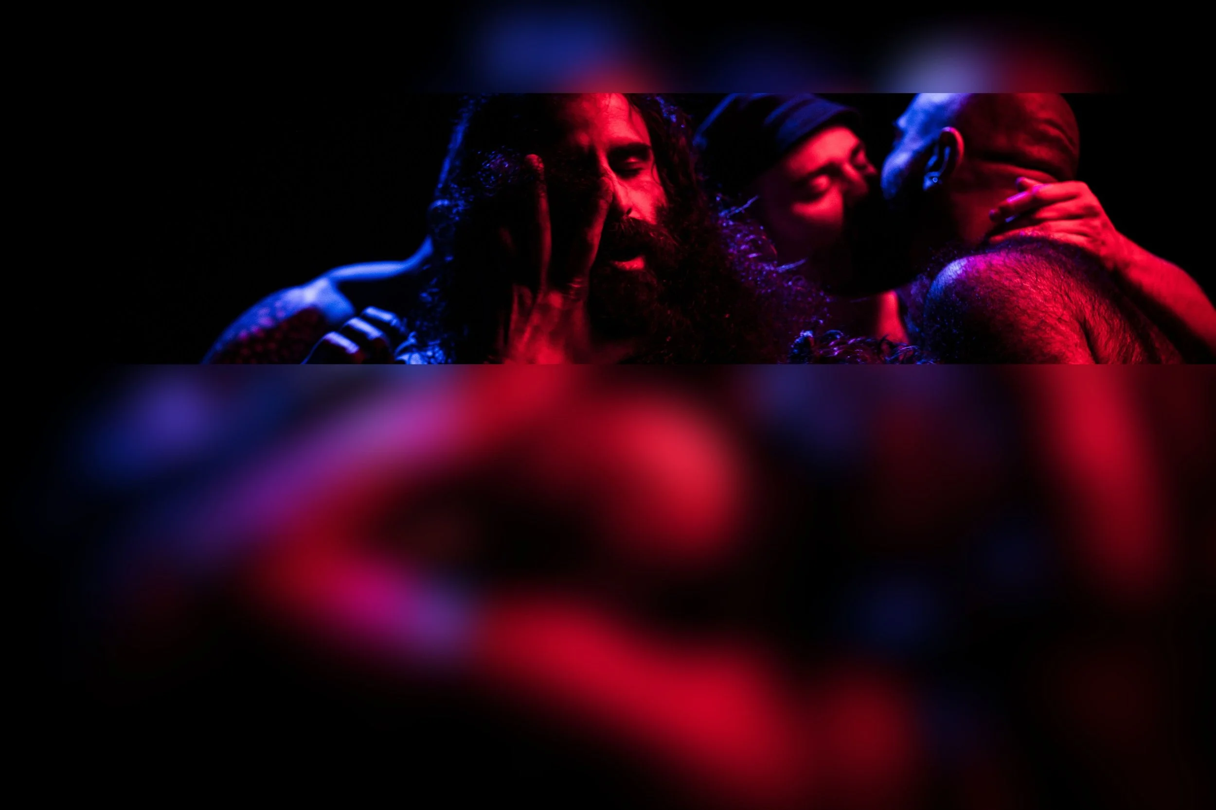 Three people in colorful lighting: one with curly hair and a beard, closed eyes and hand on face; a woman with short hair and glasses, touching face; a bald man with hand near neck, all sharing an intimate moment.