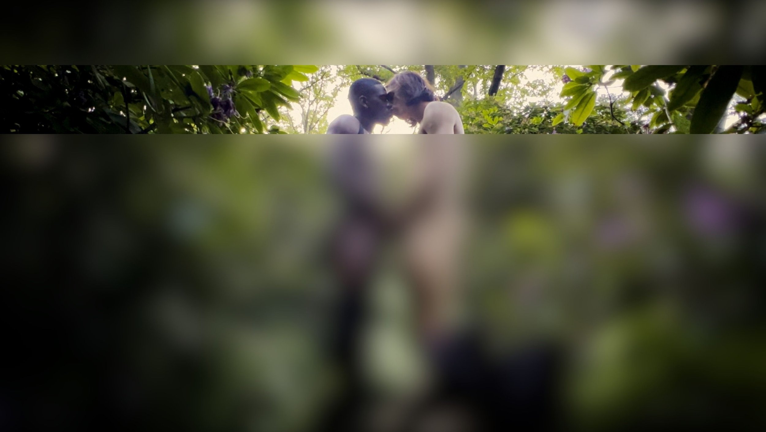 Two people with bare torsos touching foreheads in a lush green outdoor setting, surrounded by foliage.