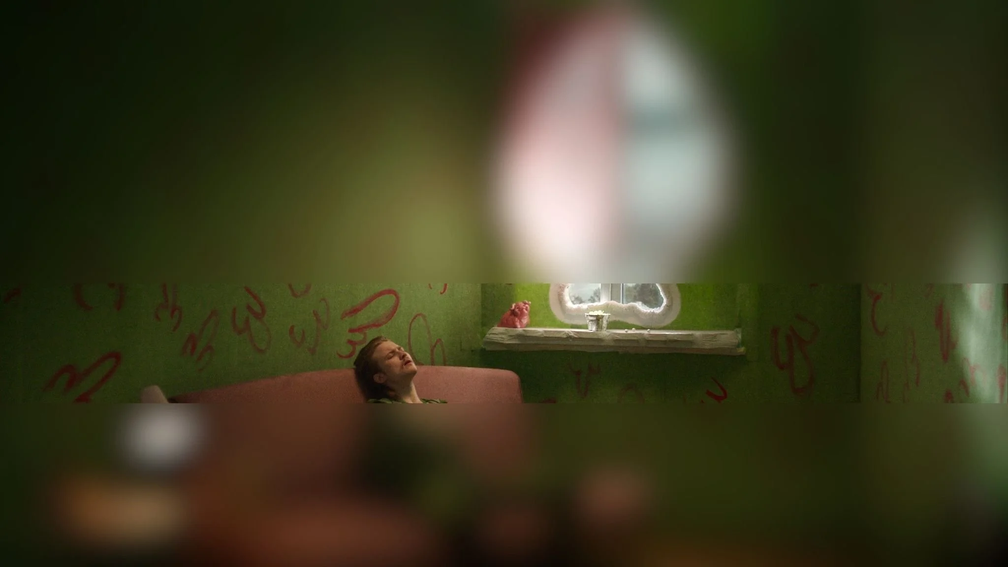 A woman sitting on a pink couch in a room with green walls, red writing or drawings on the walls, and a small window with a white frame and a flower pot on the windowsill.