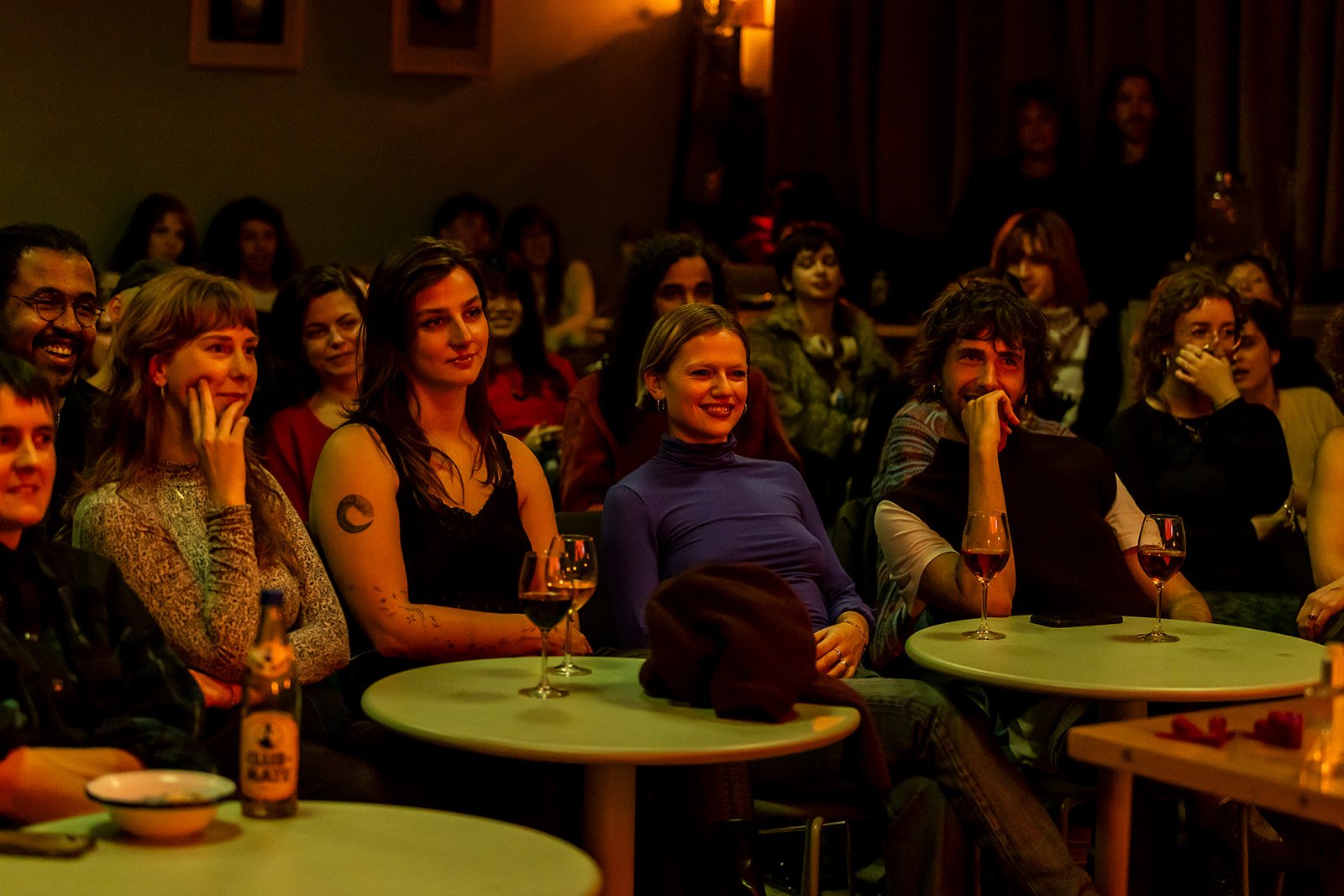 Audience at a schmutz cinema event, seated at round tables, smiling and enjoying the show in a dimly lit room.