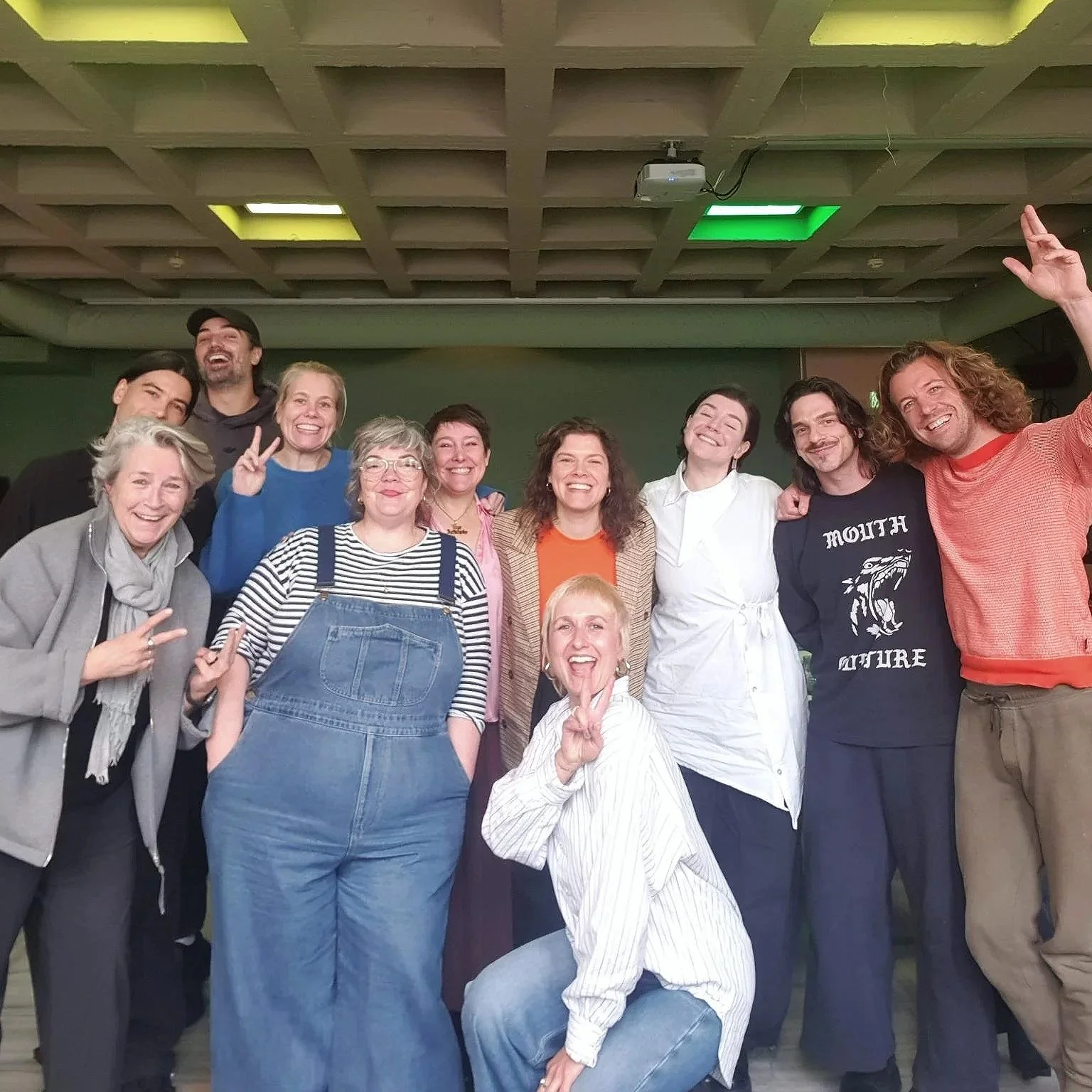 First advisory board session for Foundation House of Schmutz. A group of diverse people smiling in a room, including women, men, and one person in a white chef coat, standing under a ceiling with yellow and green lights.
